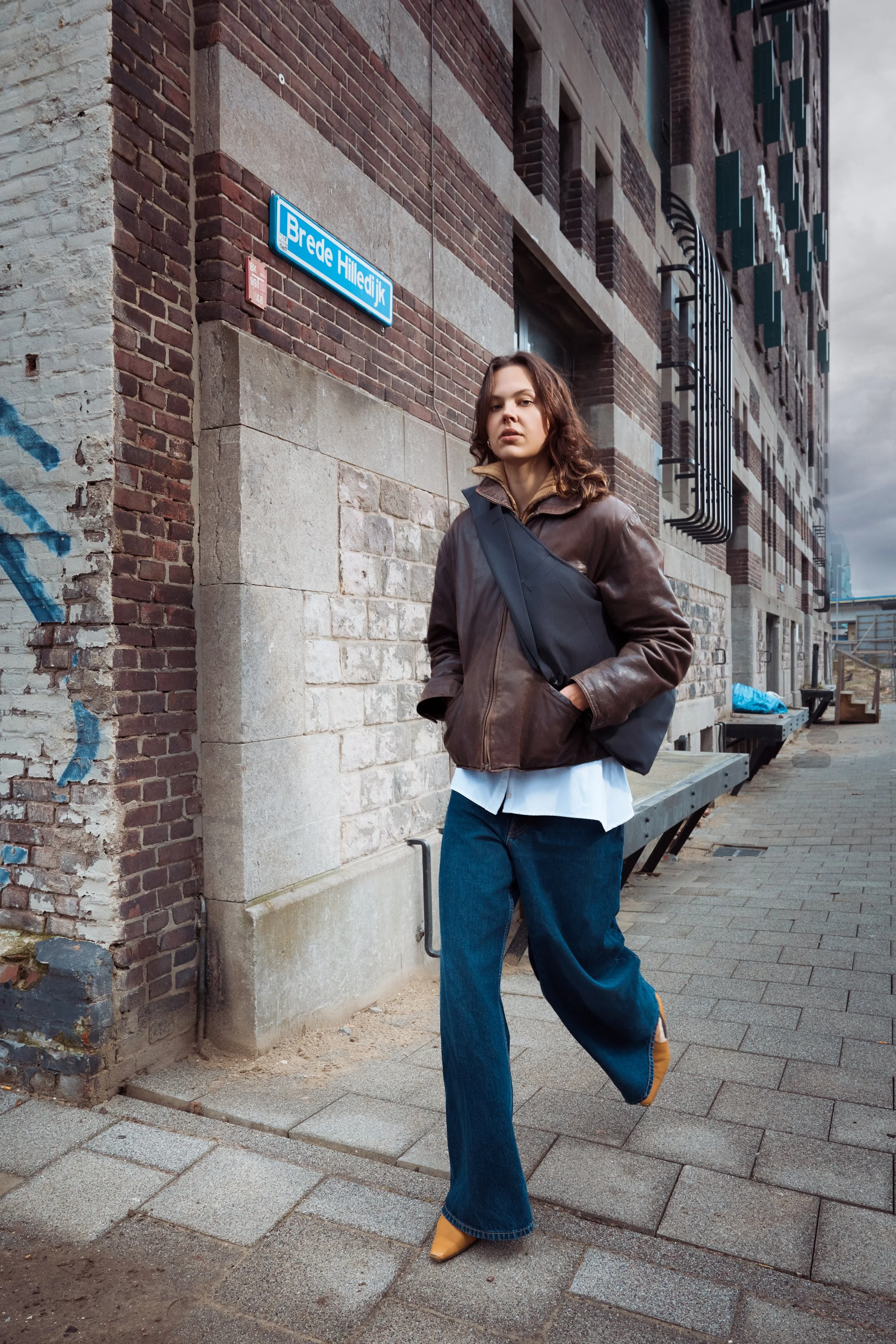 A woman walking on a city sidewalk near a brick building with a blue street sign that says 'Brede Hilledijk.' She is wearing a brown leather jacket, white shirt, blue jeans, and yellow boots, and is carrying a black shoulder bag.