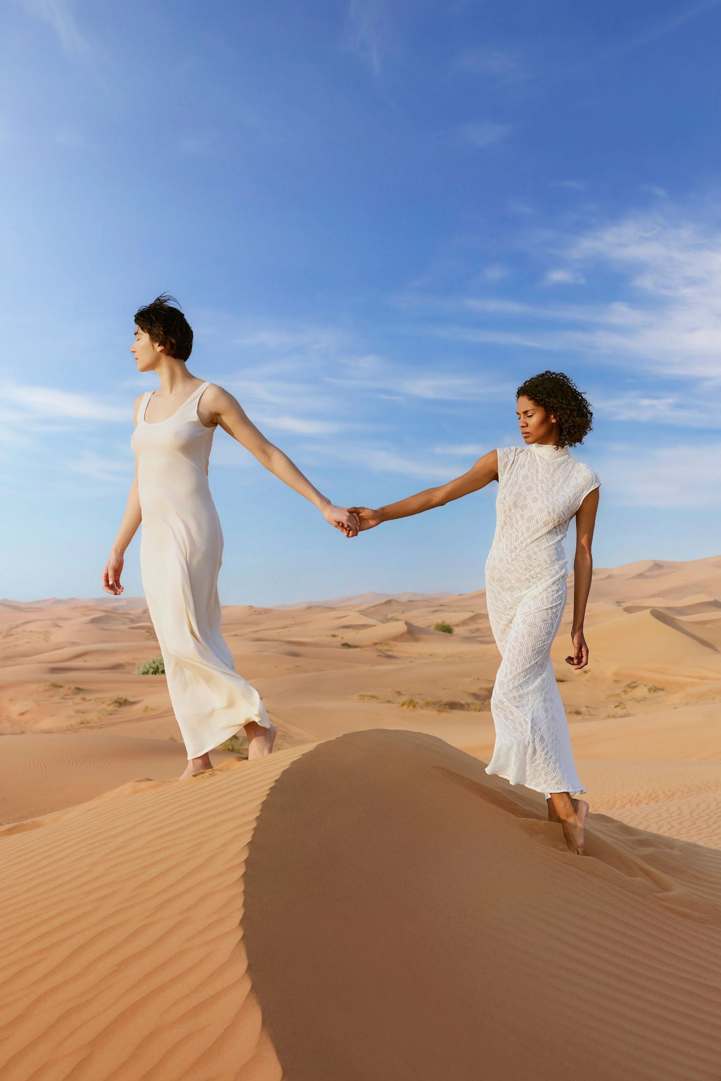 Two women in white dresses walking in a desert, holding hands, with sand dunes and blue sky in the background.