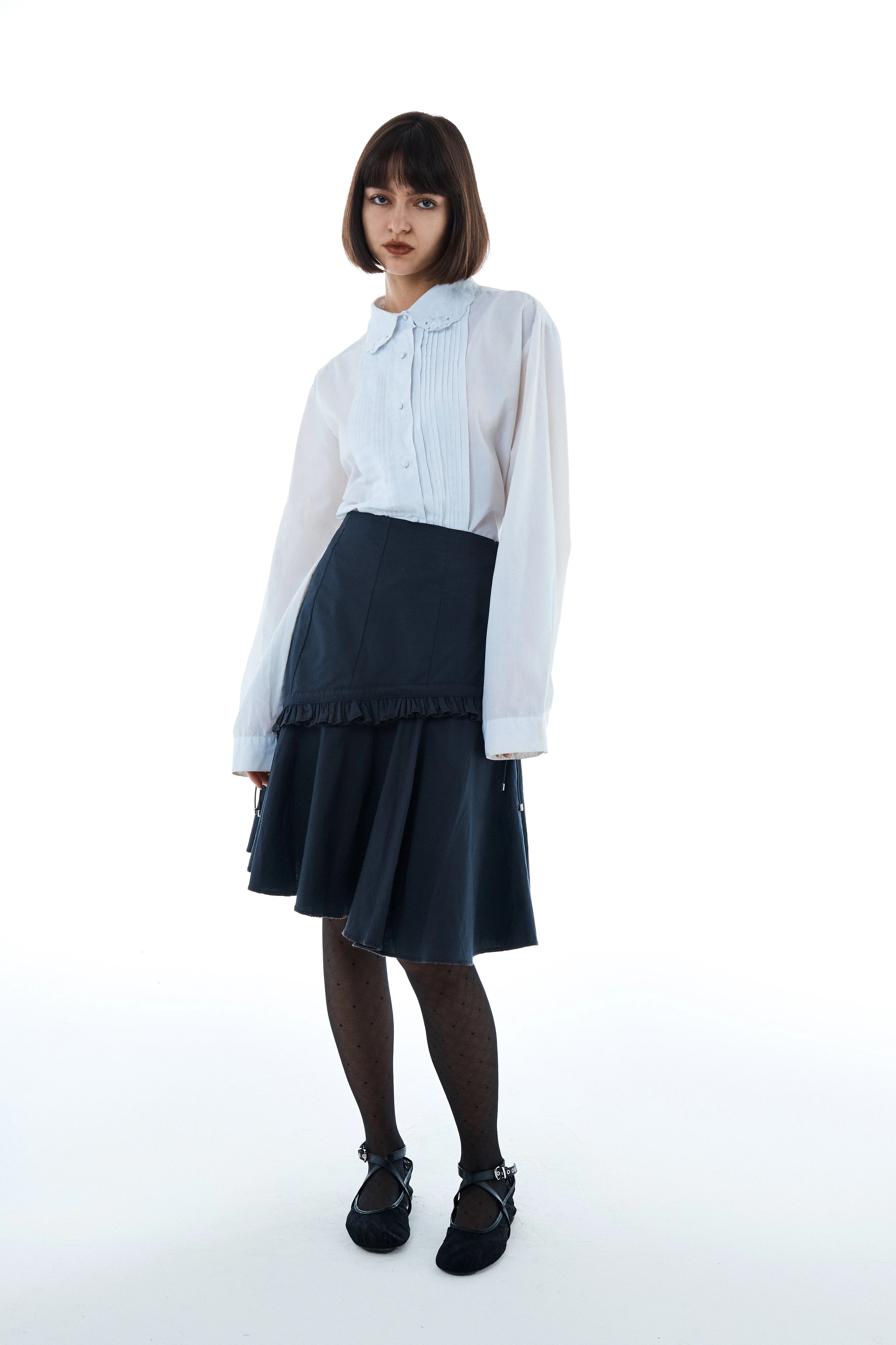 Young woman with dark hair wearing a white blouse, navy skirt with ruffle detail, black patterned tights, and black shoes, standing against a white background.