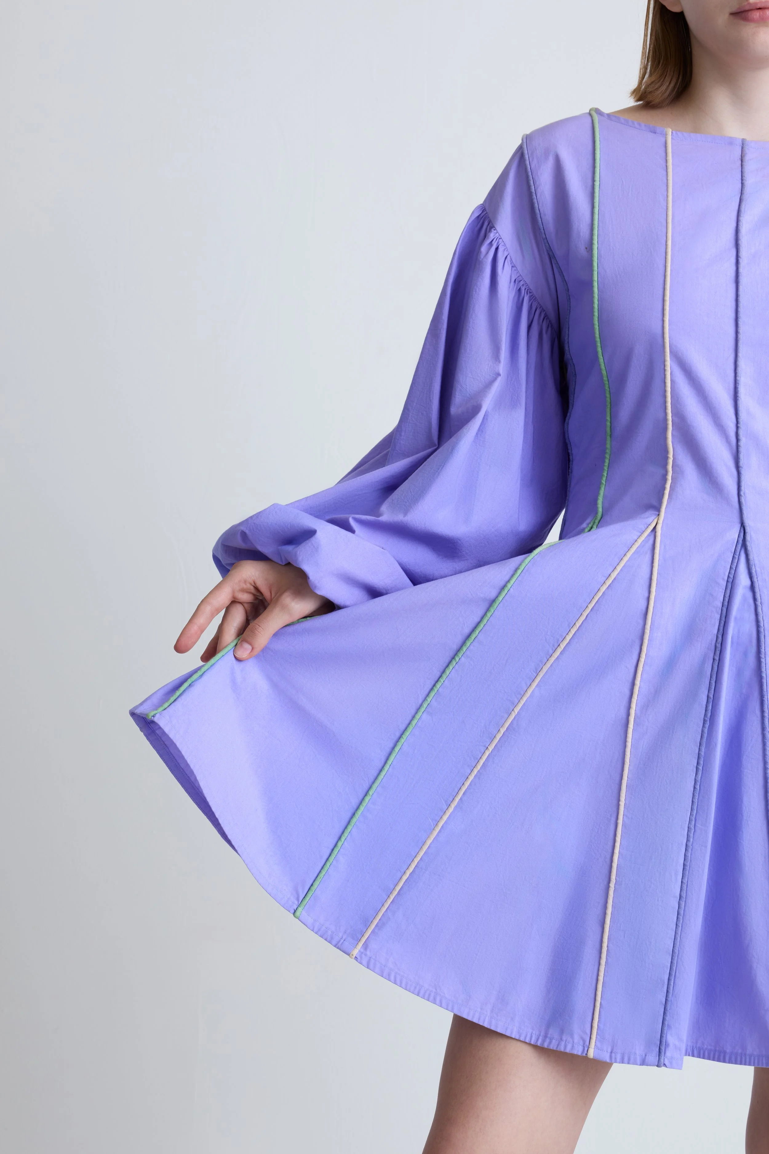 A woman wearing a lavender dress with colorful stitching details, holding the bottom of her dress to show its full shape.