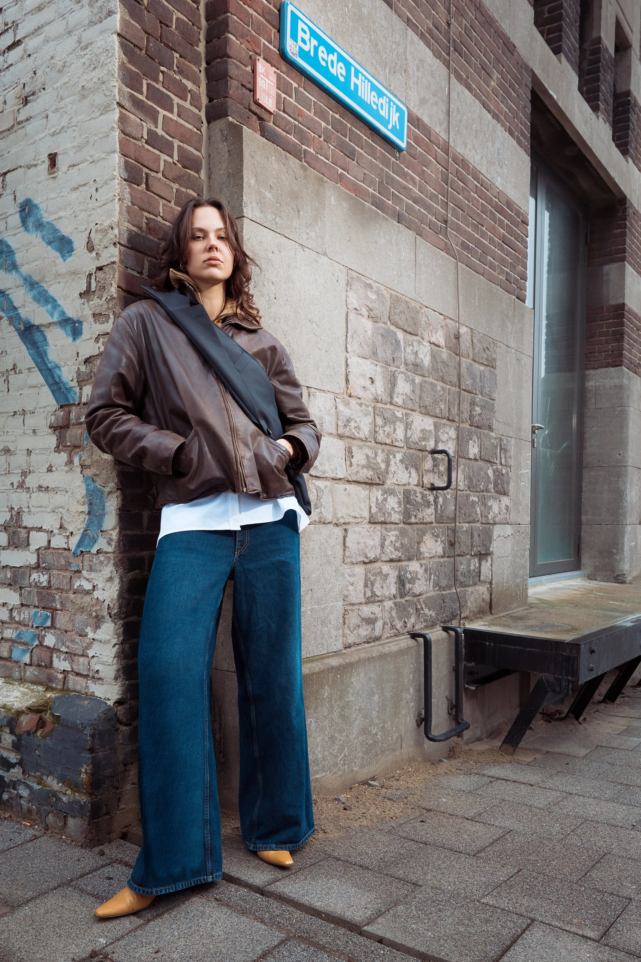 A woman standing against a brick wall on a city sidewalk, wearing a brown leather jacket, white shirt, wide-leg jeans, and tan boots. She has a black crossbody bag and looks serious. A blue street sign reads Brede Hilledijk.