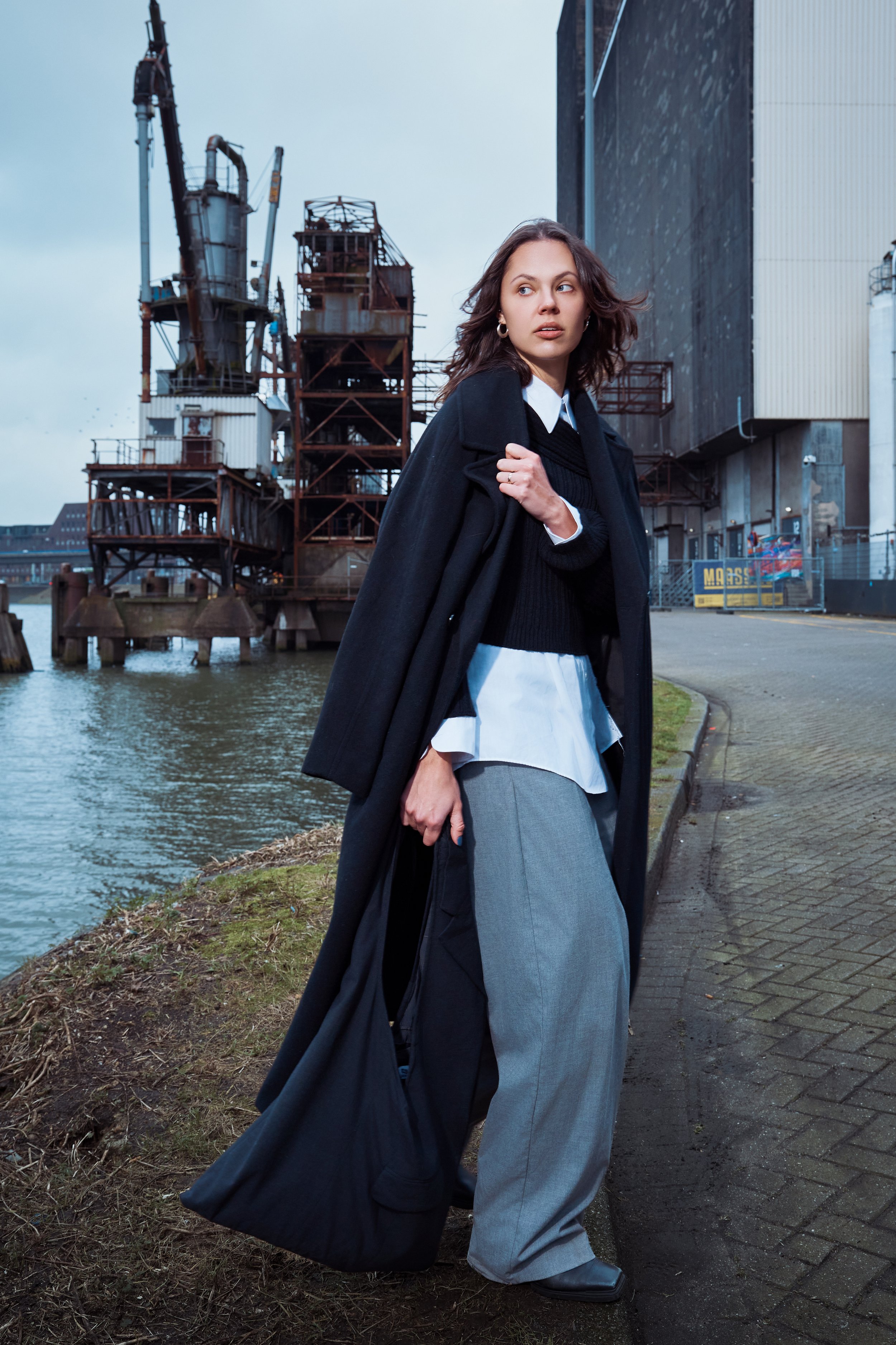 A woman dressed in fashionable layered winter clothing standing near a body of water with industrial structures in the background.
