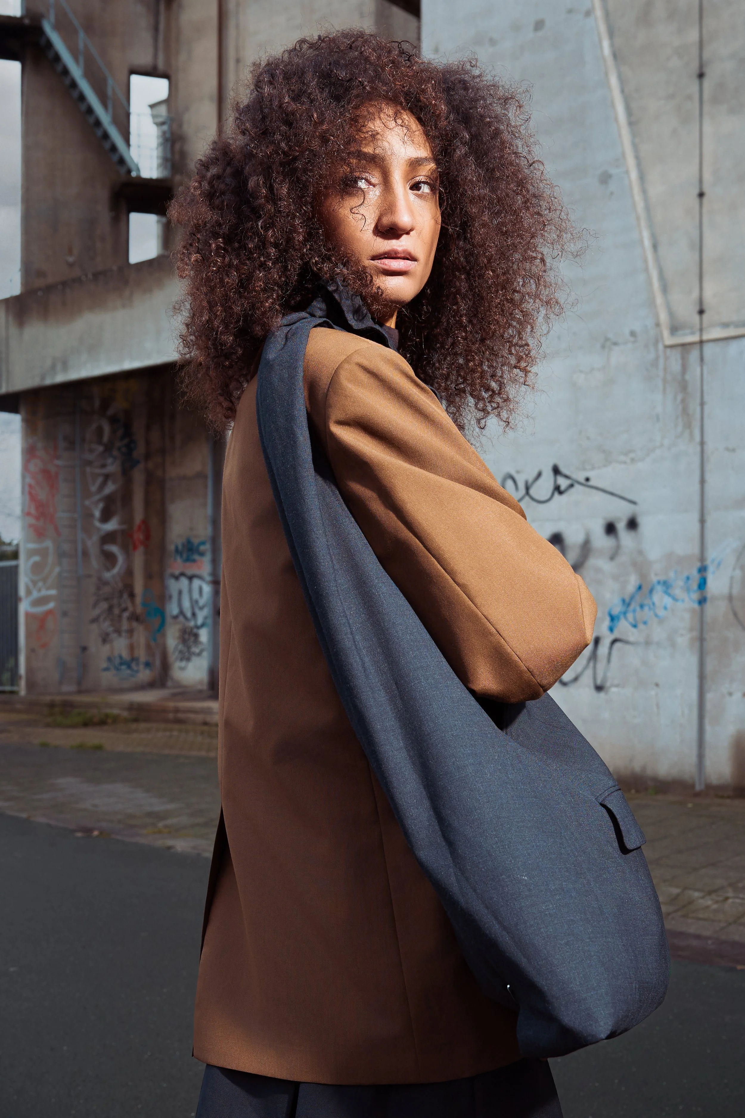 Young woman with curly hair standing outdoors in an urban setting, wearing a brown and gray coat.