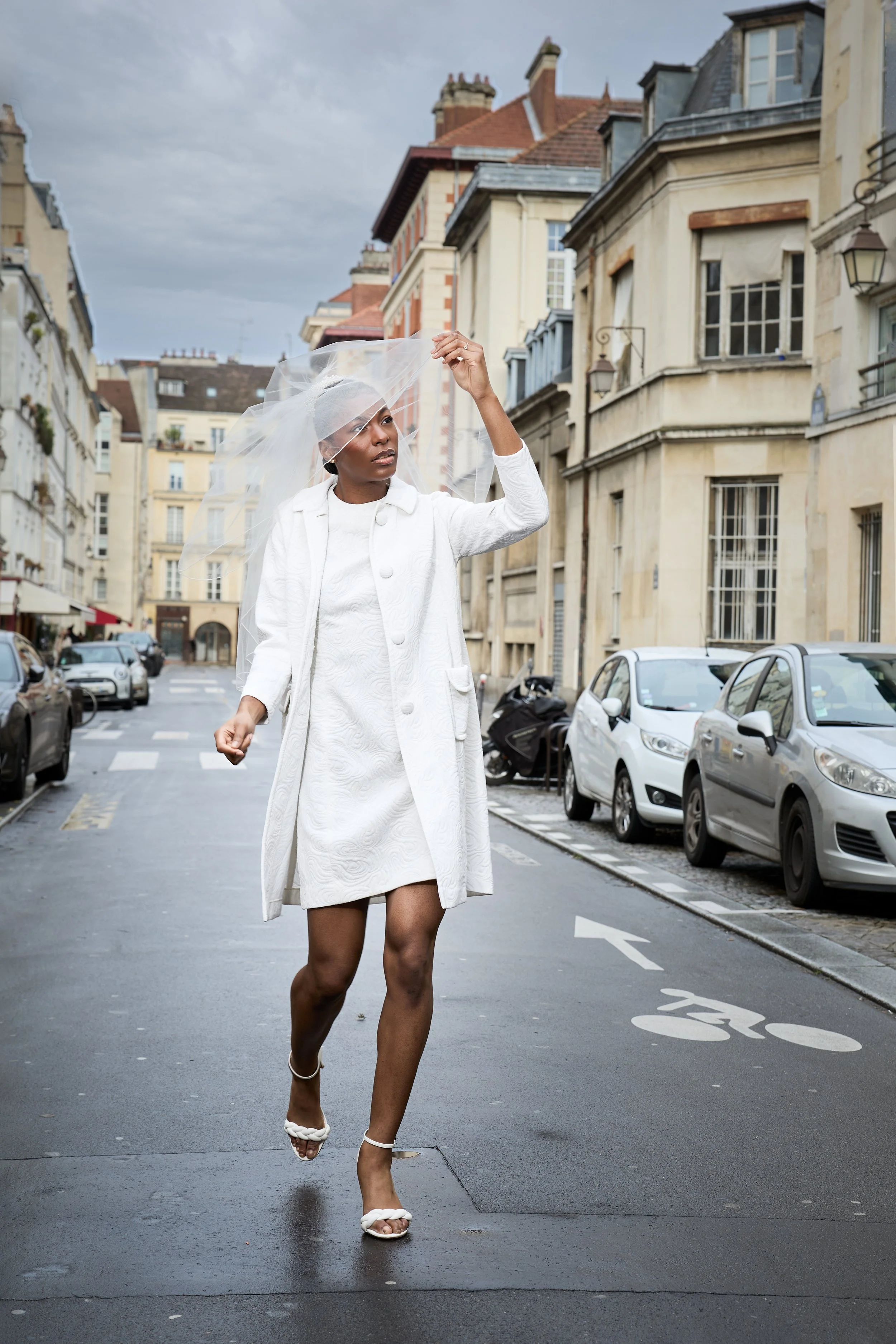 Woman dressed in white, walking on a city street, holding a wedding veil. She is wearing a white coat, dress, and heels.