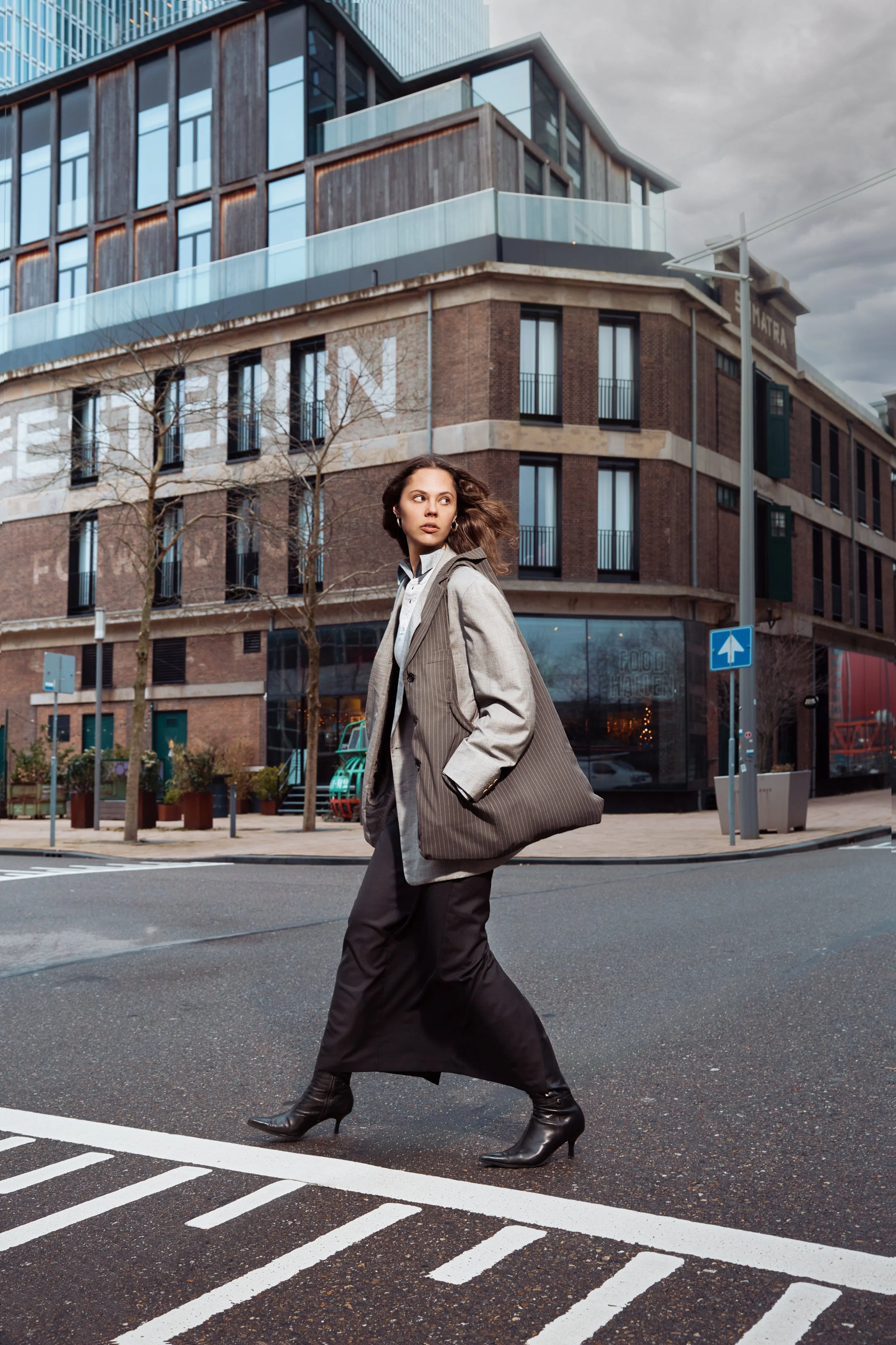 A woman walking across an urban street intersection during daytime, wearing a blazer, long skirt, and high-heeled boots, with modern buildings in the background.