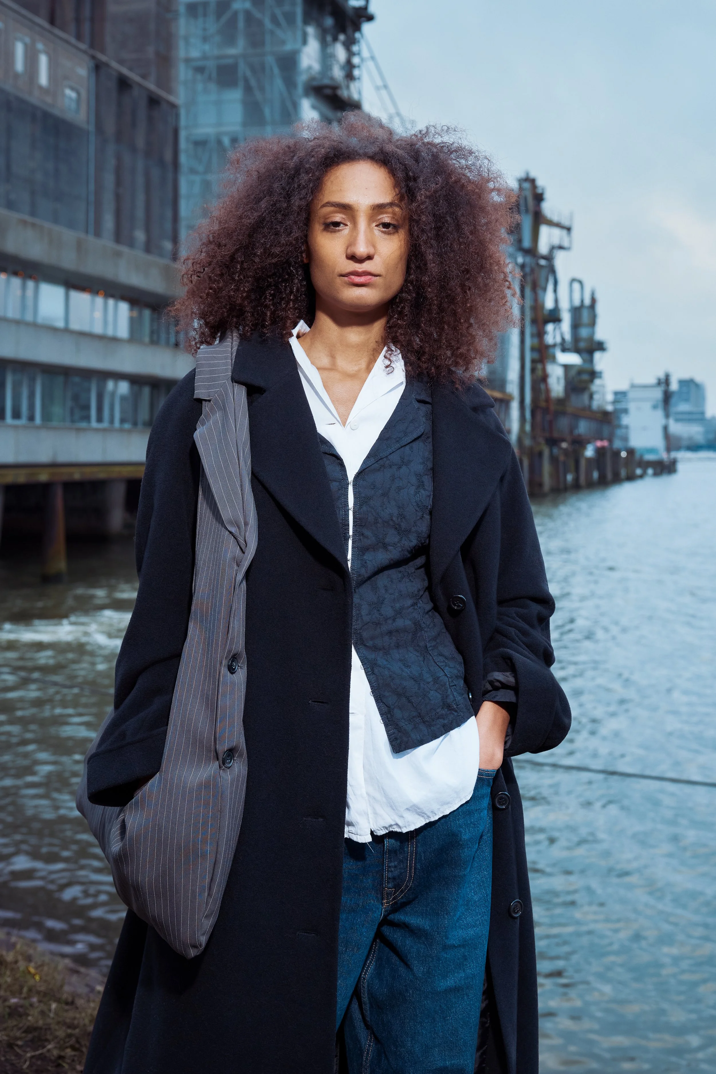 A woman with curly hair standing by water with industrial buildings in the background, wearing a dark coat, white shirt, and carrying a striped bag.