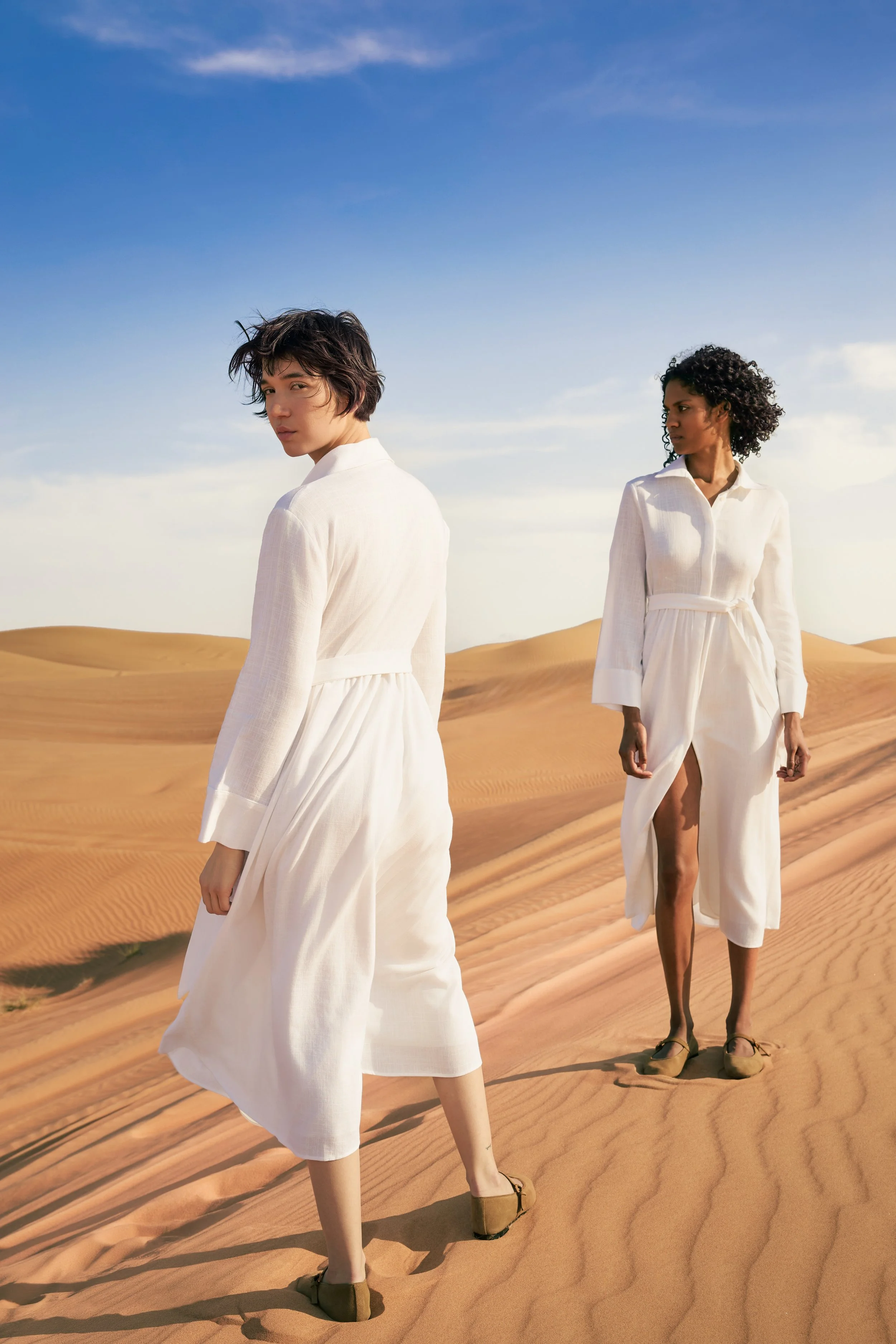 Two women wearing white dresses walk on sand dunes in a desert landscape with blue sky.