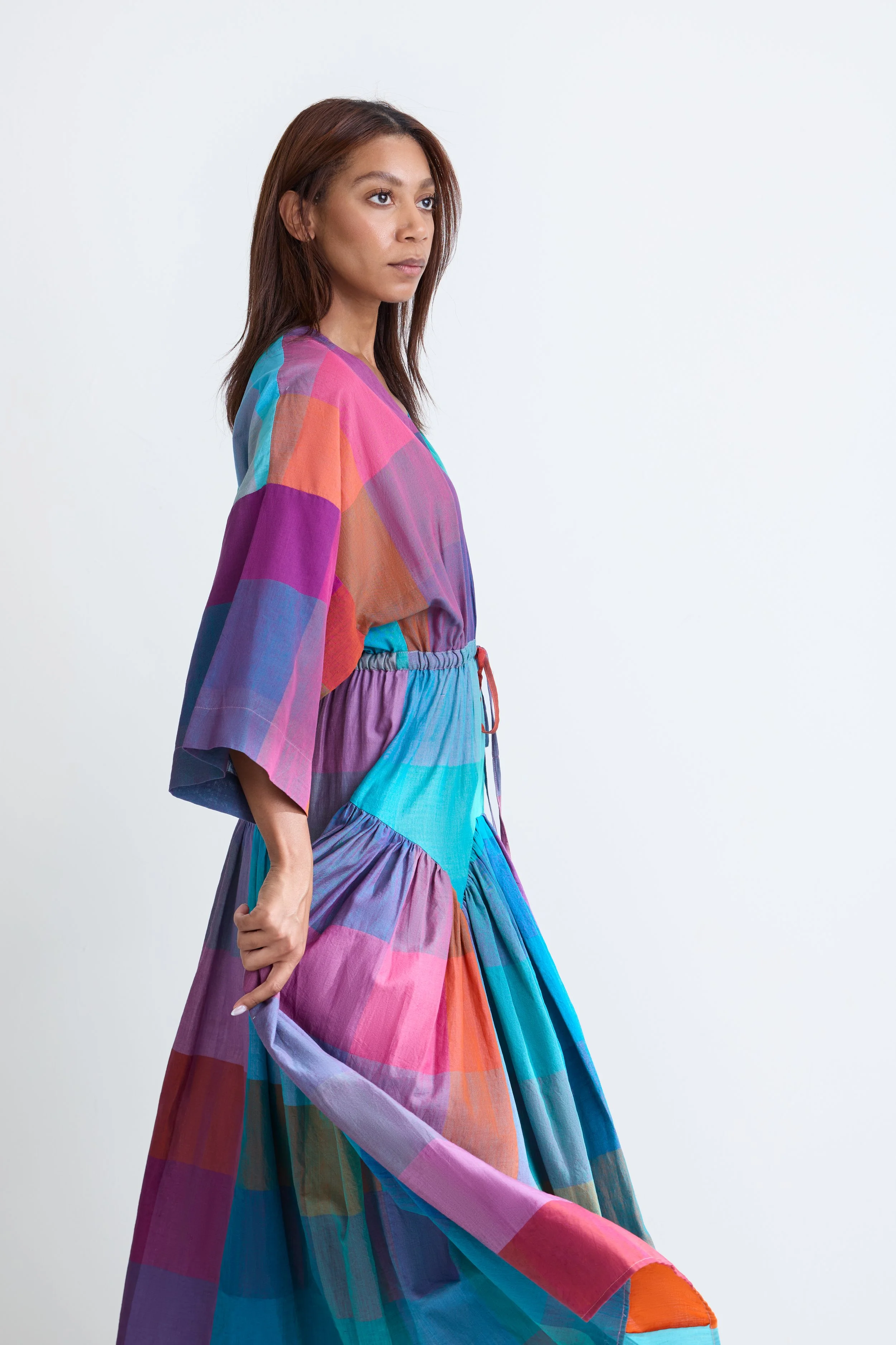 A woman with brown hair wearing a colorful, patterned, multi-patch dress standing against a plain white background.