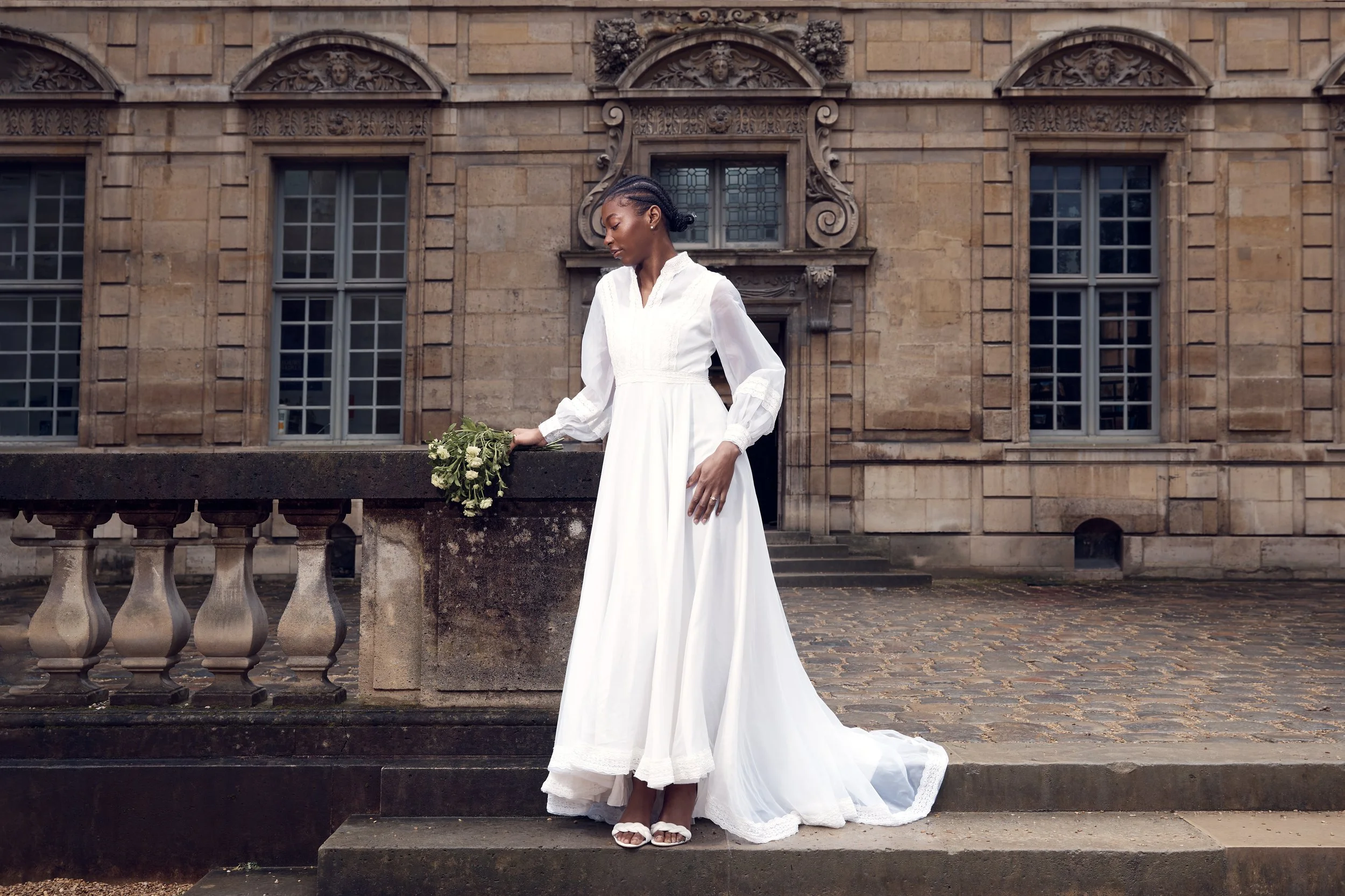A woman in a long white wedding dress holding a bouquet of flowers stands outdoors on a stone patio in front of an ornate historic stone building.