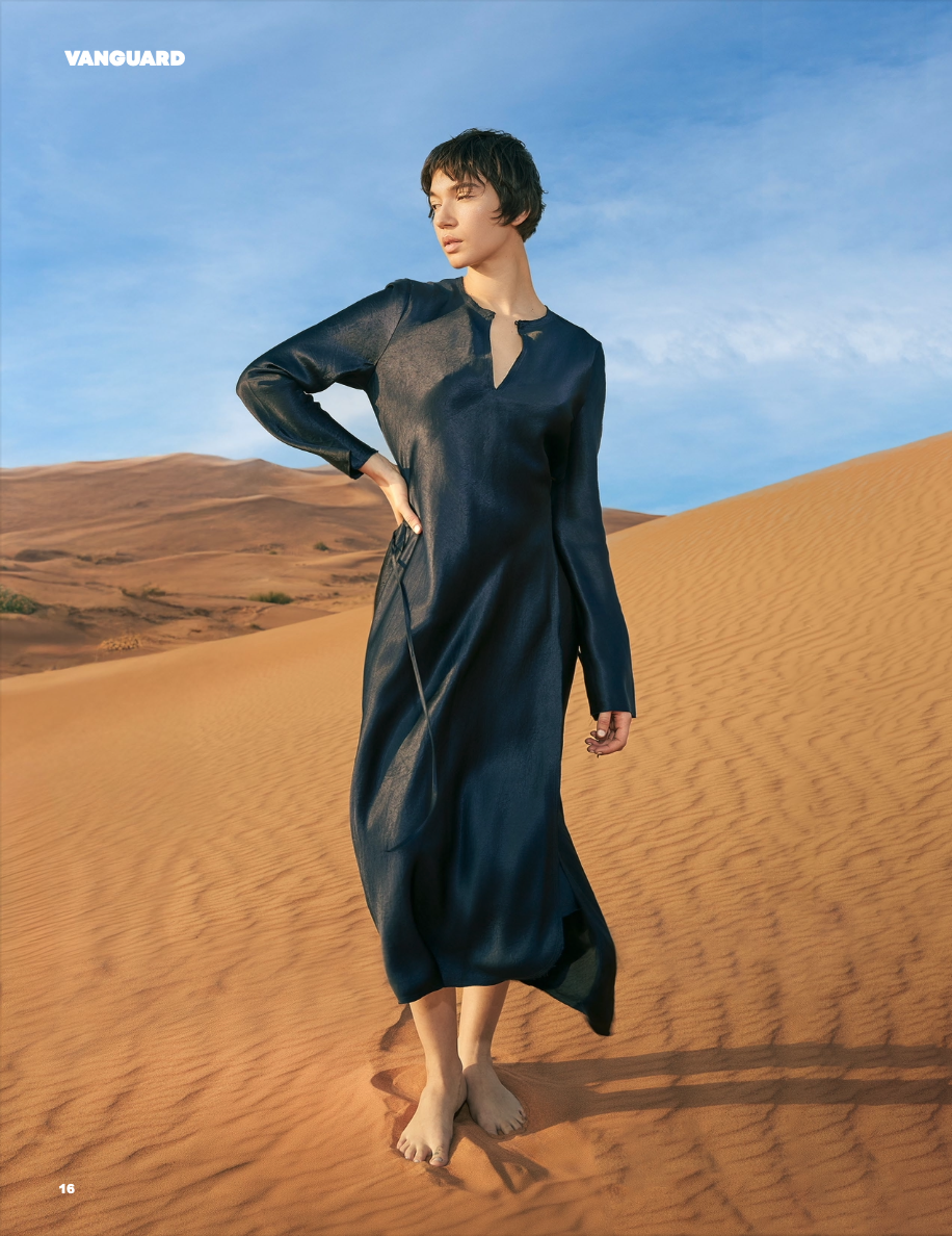 A woman in a long, dark dress standing barefoot on sand dunes in a desert landscape with a partly cloudy sky.