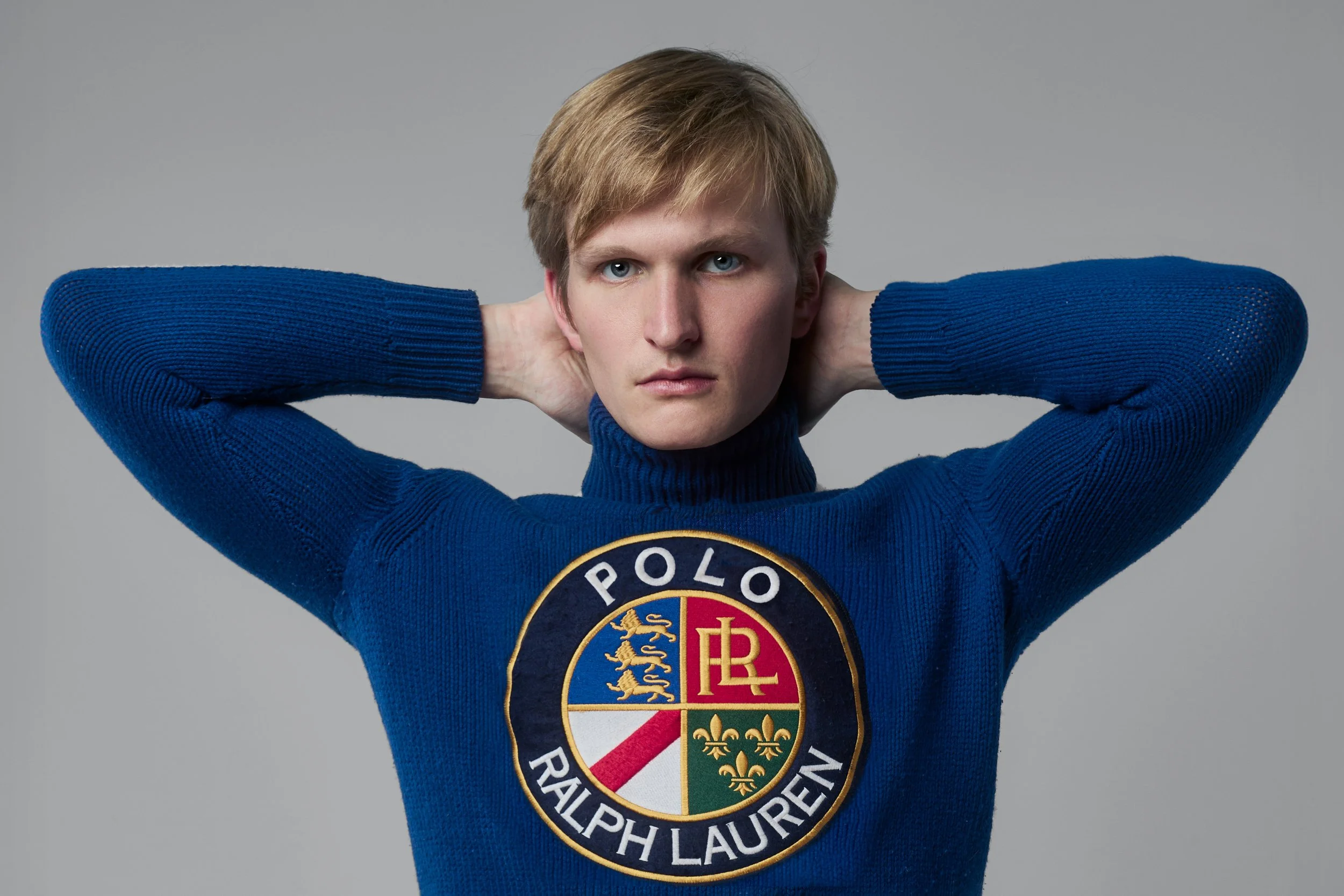 Young man with blonde hair wearing a blue sweater that features the Polo Ralph Lauren logo, posing with hands behind his head against a neutral background.