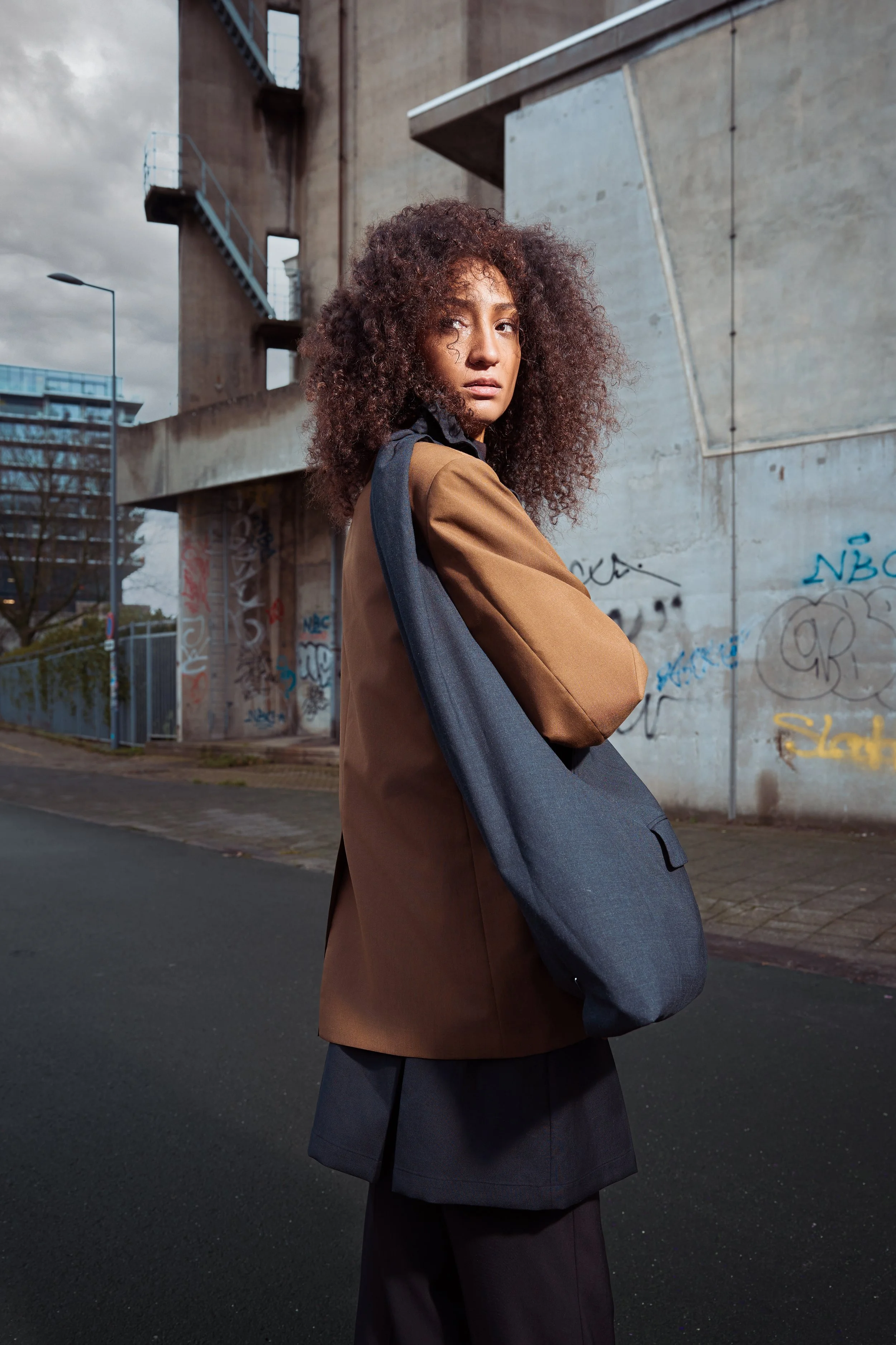 A woman with curly hair stands on a city street, wearing a brown blazer, black pants, and carrying a large gray bag, with urban architecture and graffiti in the background.