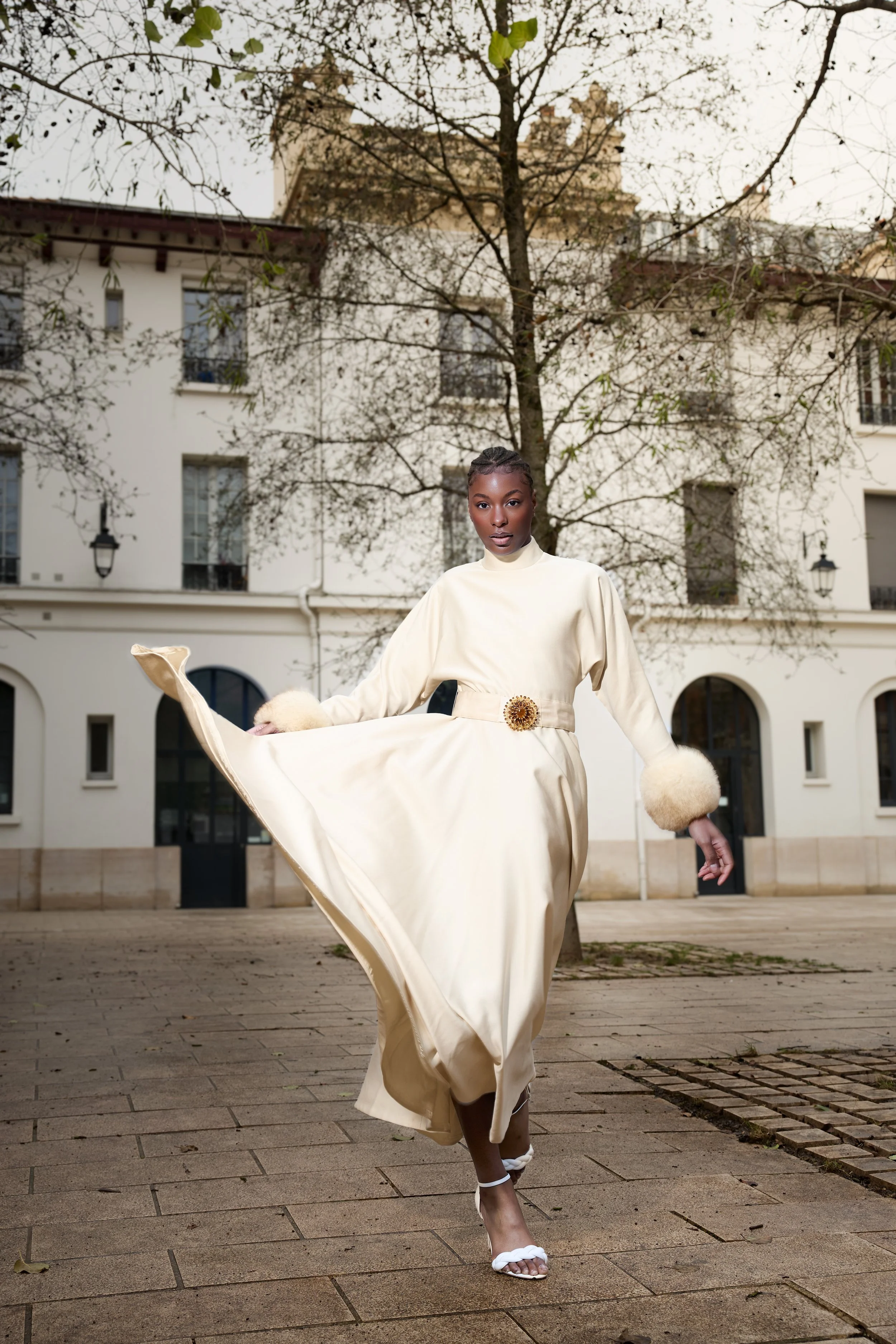 A woman in an elegant cream-colored dress with puffed sleeves and fluffy cuffs, walking outdoors on a paved area, with buildings and a tree in the background.