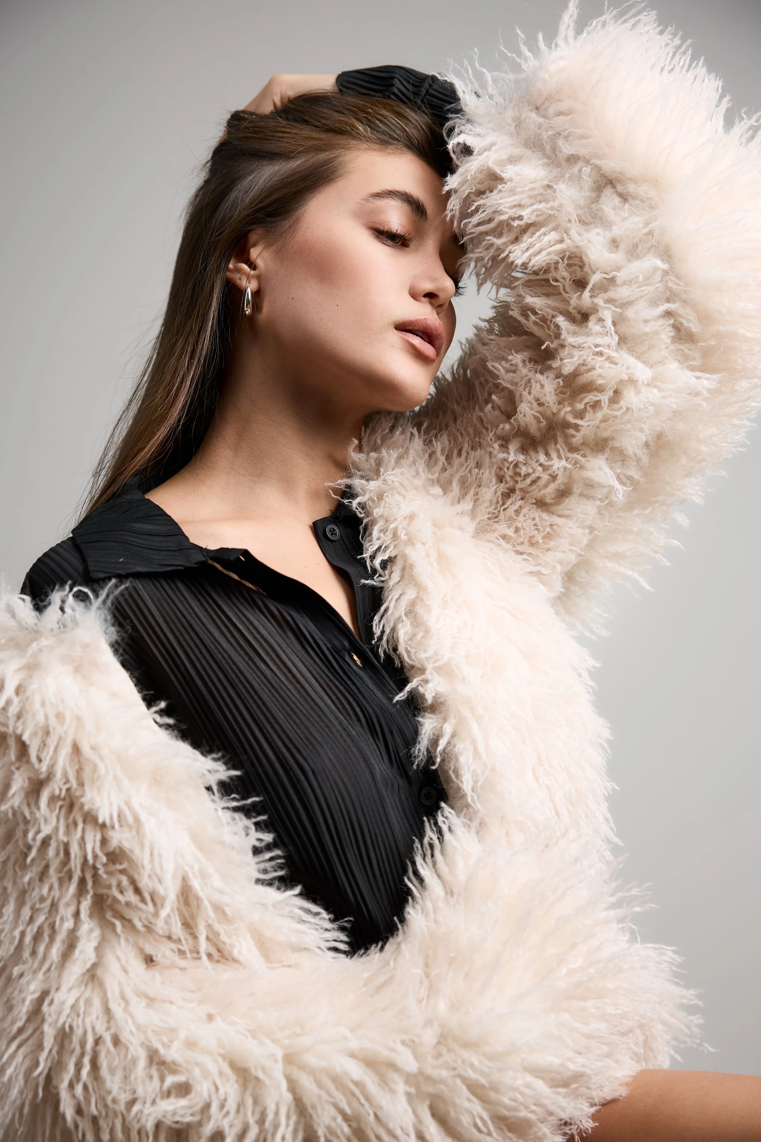 A woman with long brown hair wearing earrings poses with a fluffy cream-colored jacket over a black pleated shirt.