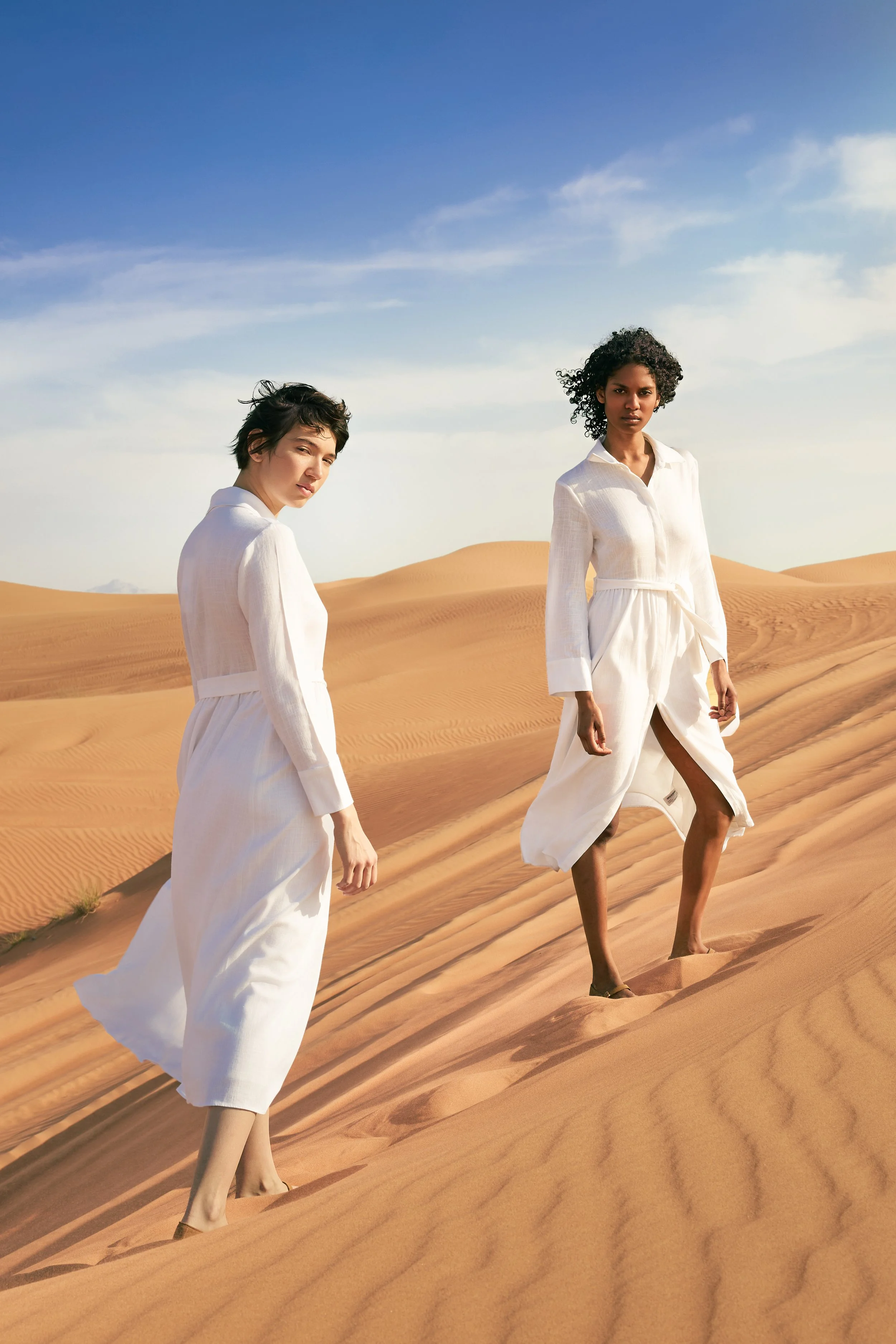 Two women in white dresses walking on sand dunes in a desert under a blue sky.