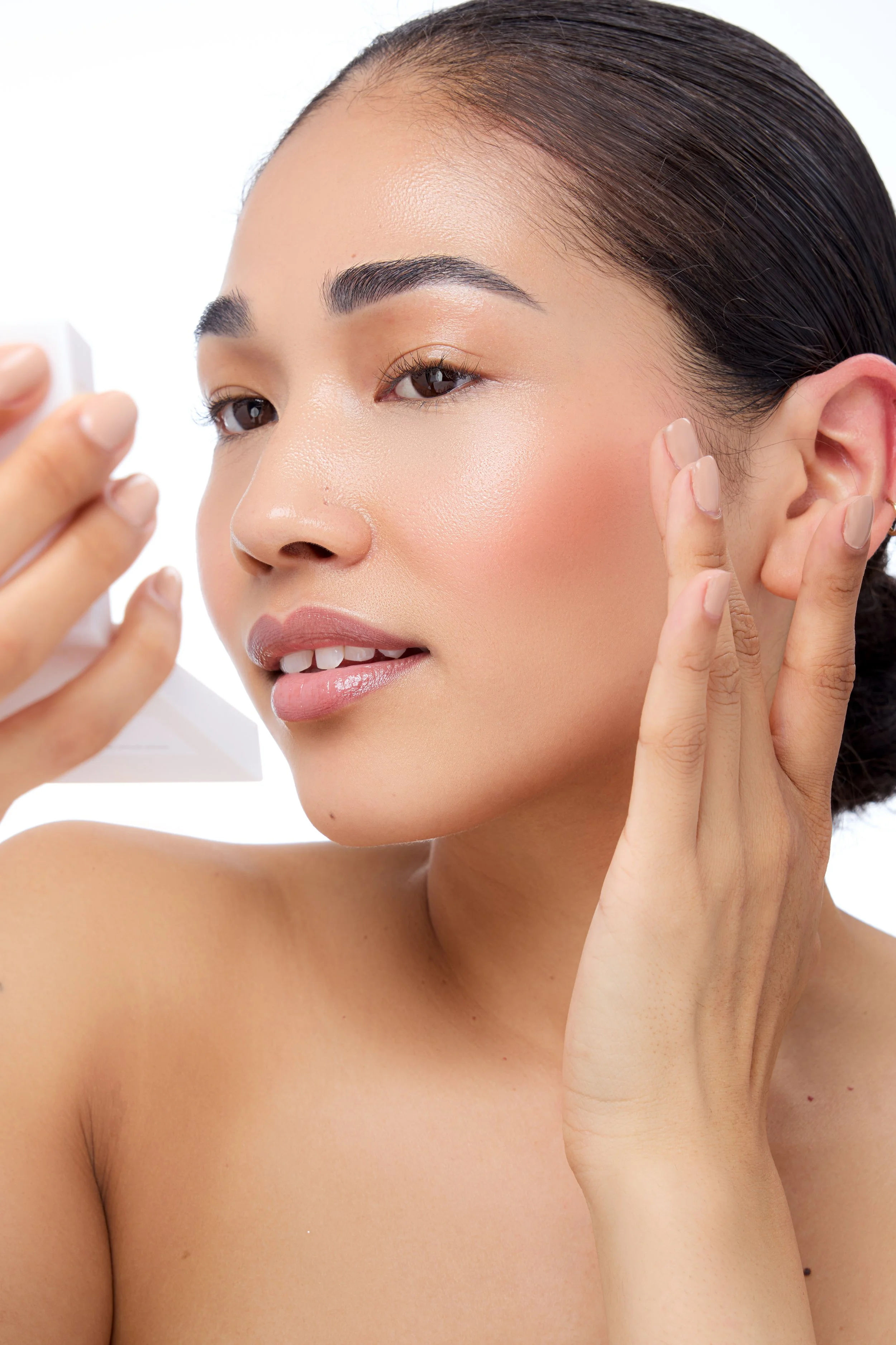 A woman with flawless skin is applying skincare or makeup to her face, holding a small container in one hand and gently touching her cheek with the other.