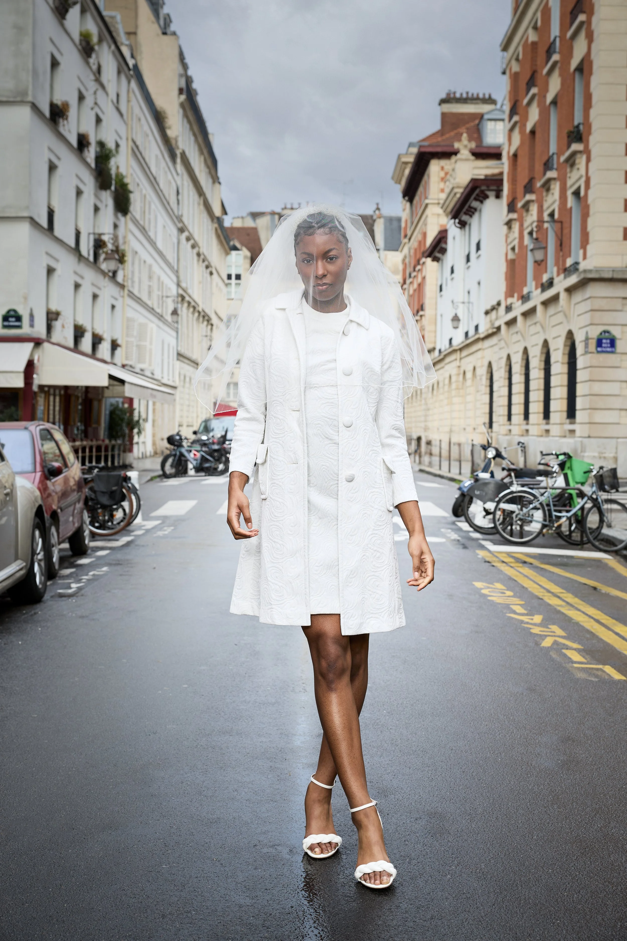 A woman walking down a city street wearing a white trench coat, white high heels, and a veil, with buildings and parked bicycles in the background.