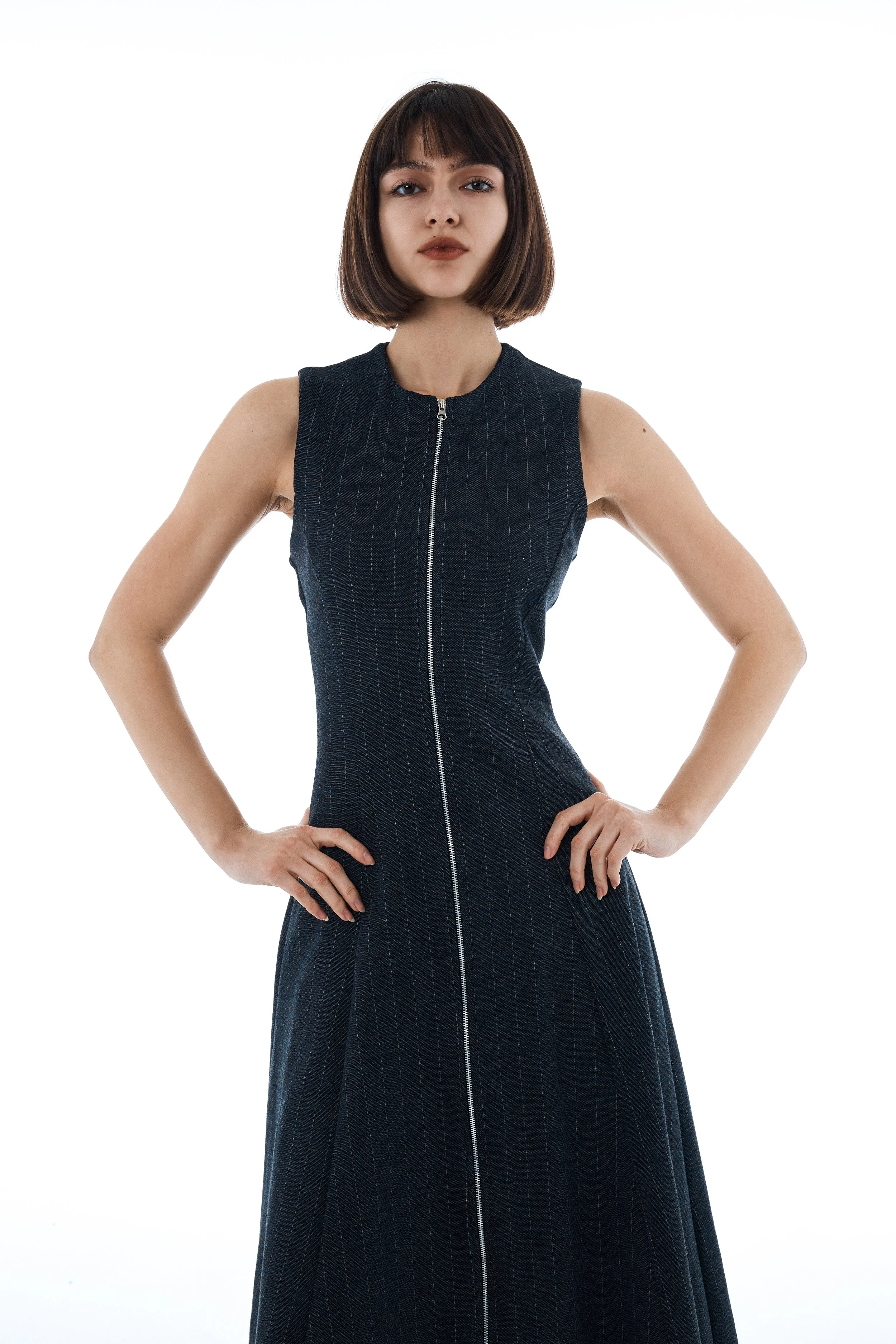 Woman with short brown hair wearing a sleeveless black dress with a front zipper, standing with hands on hips, against a plain white background.