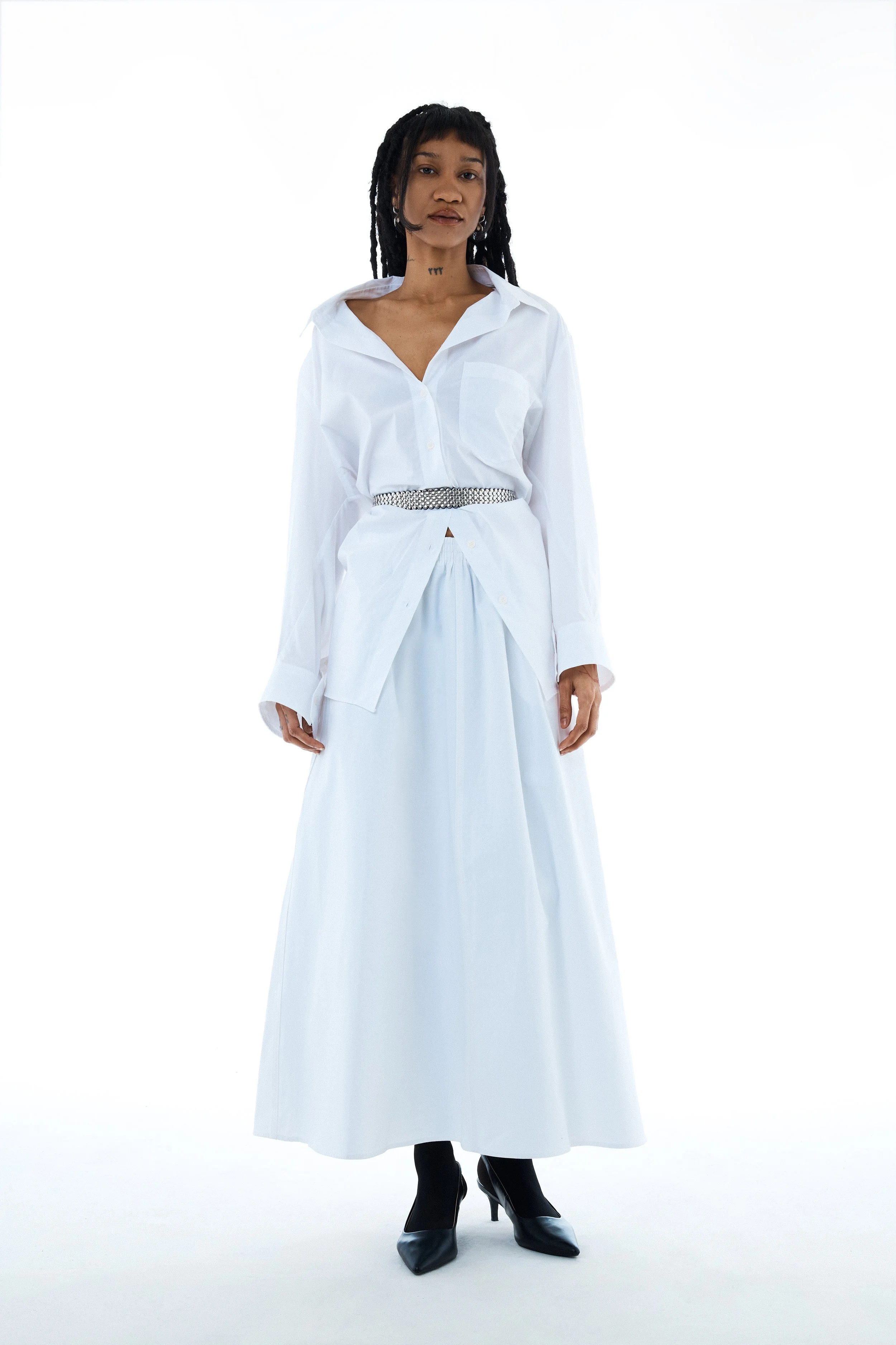 Woman in a white shirt, long white skirt, black heels, standing against a white background.