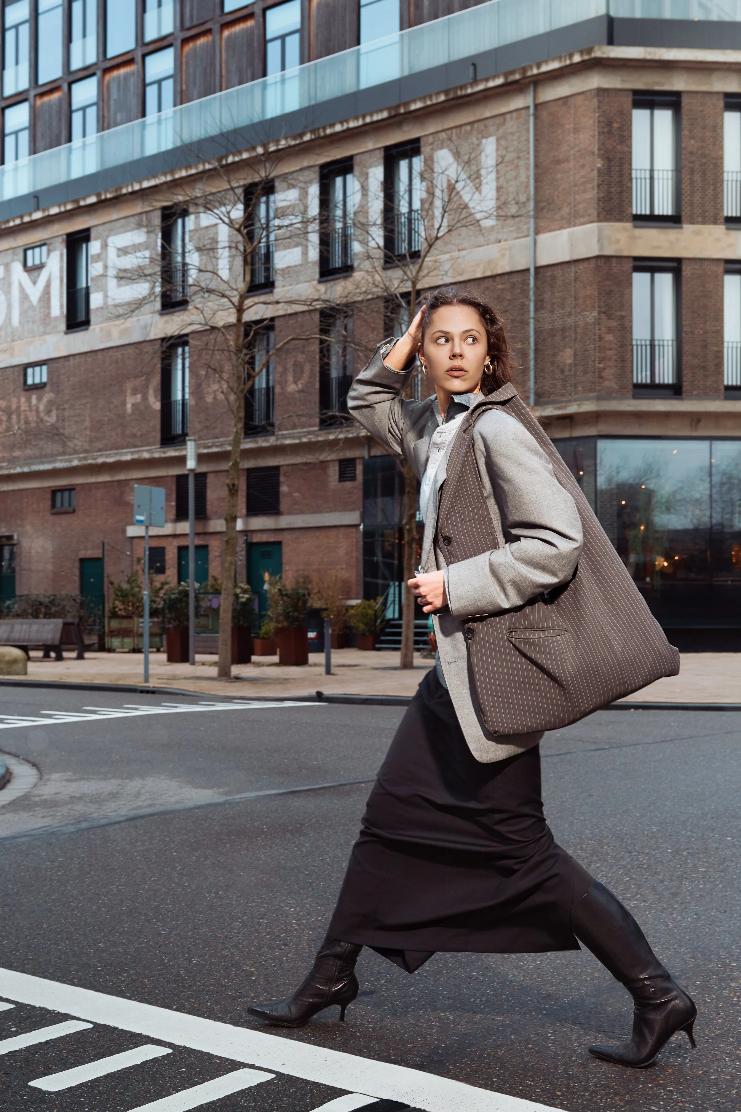 Fashionably dressed woman crossing the street in an urban area, wearing a gray blazer, long black skirt, and black high-heeled boots, with a large brown striped bag, in front of modern buildings.