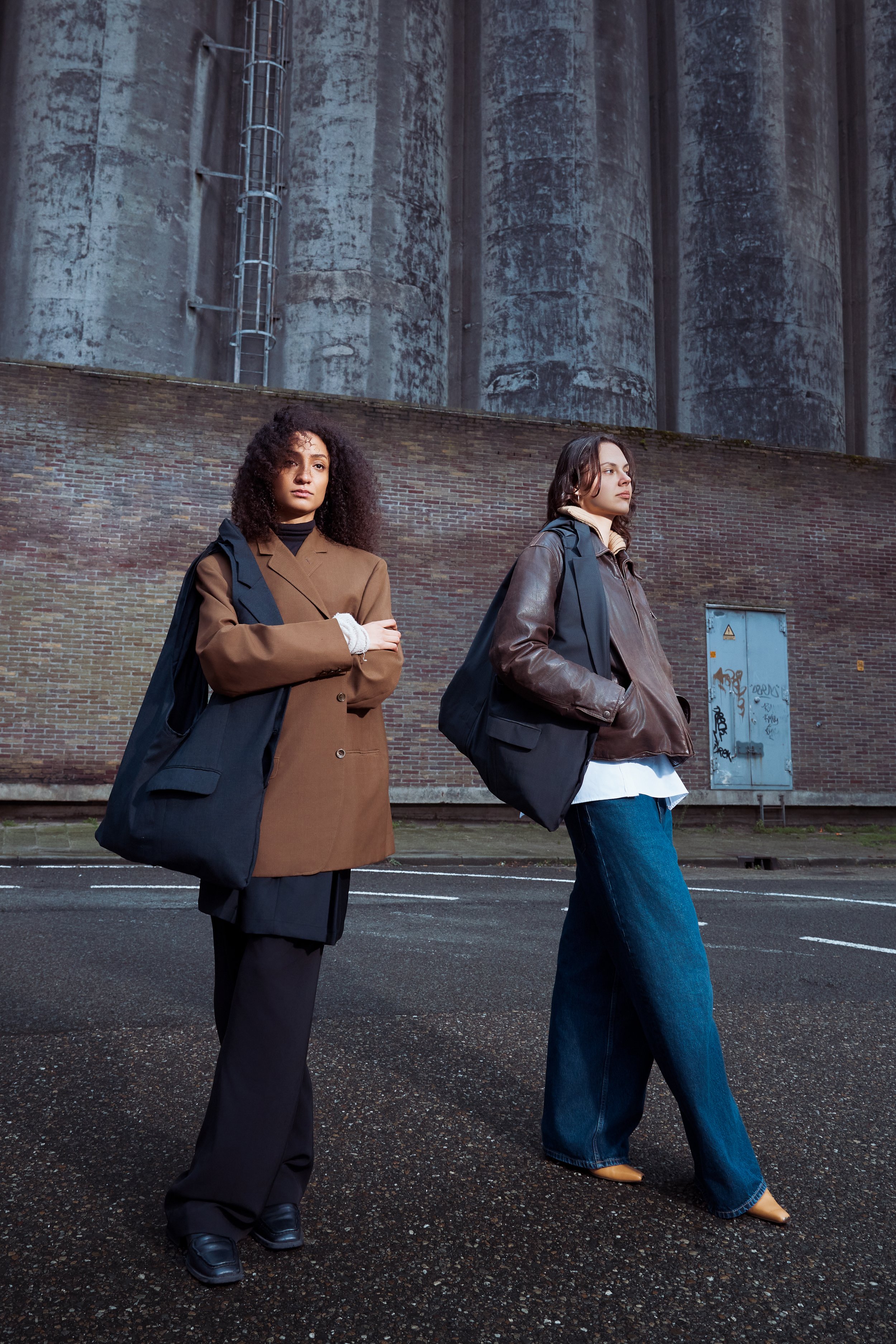 Two young women standing on a city street at night, dressed in stylish casual coats and jeans, carrying black bags, against an industrial background with large silos and graffiti.
