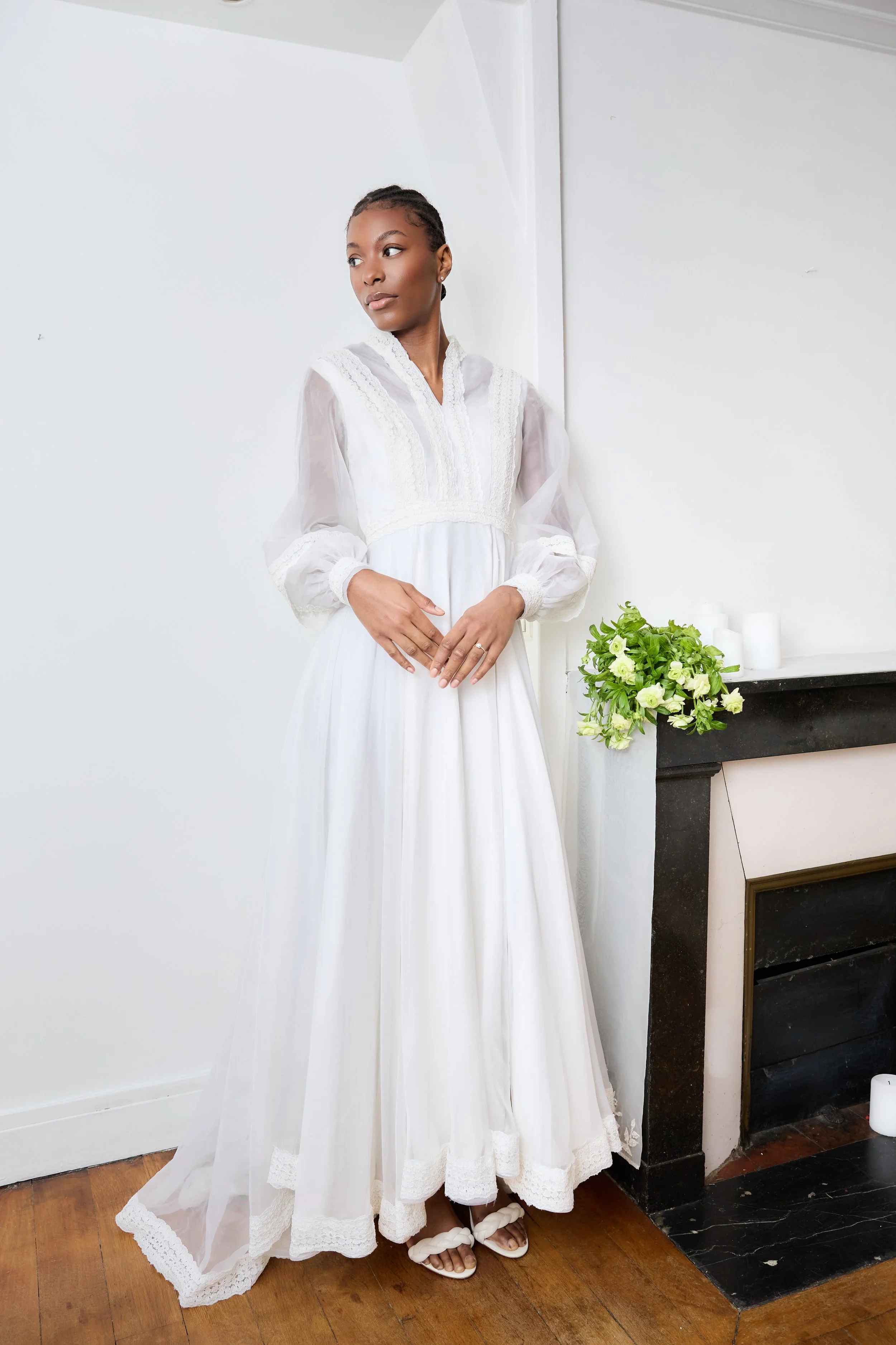 A woman in a white dress standing indoors near a black fireplace with white candles and a bouquet of green and white flowers.