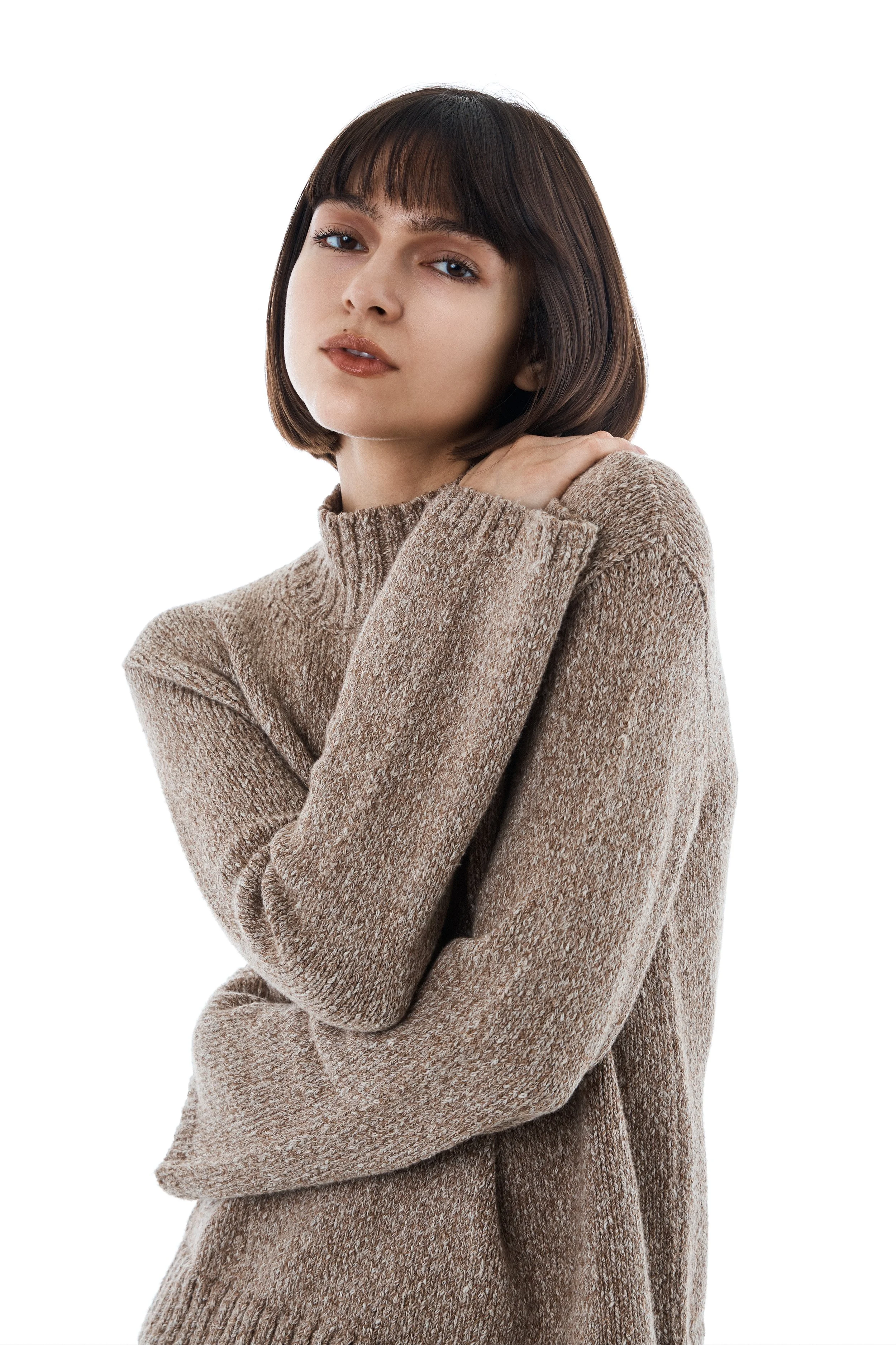 Young woman with short brown hair, wearing a beige sweater, posing with one hand on her shoulder against a white background.