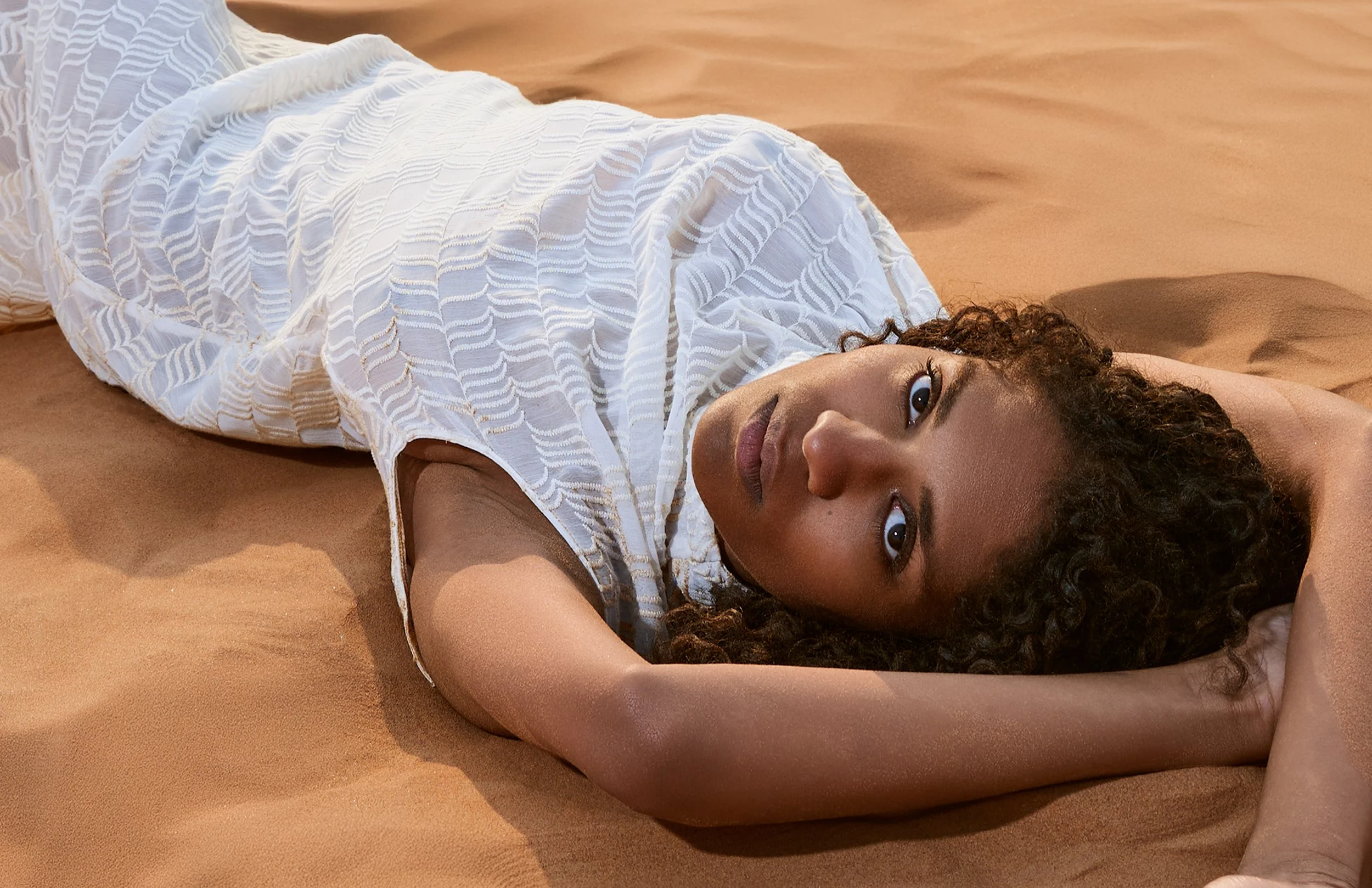 A woman with curly hair lying on her back on a sandy surface, wearing a white textured dress, with her head turned towards the camera, gazing directly.
