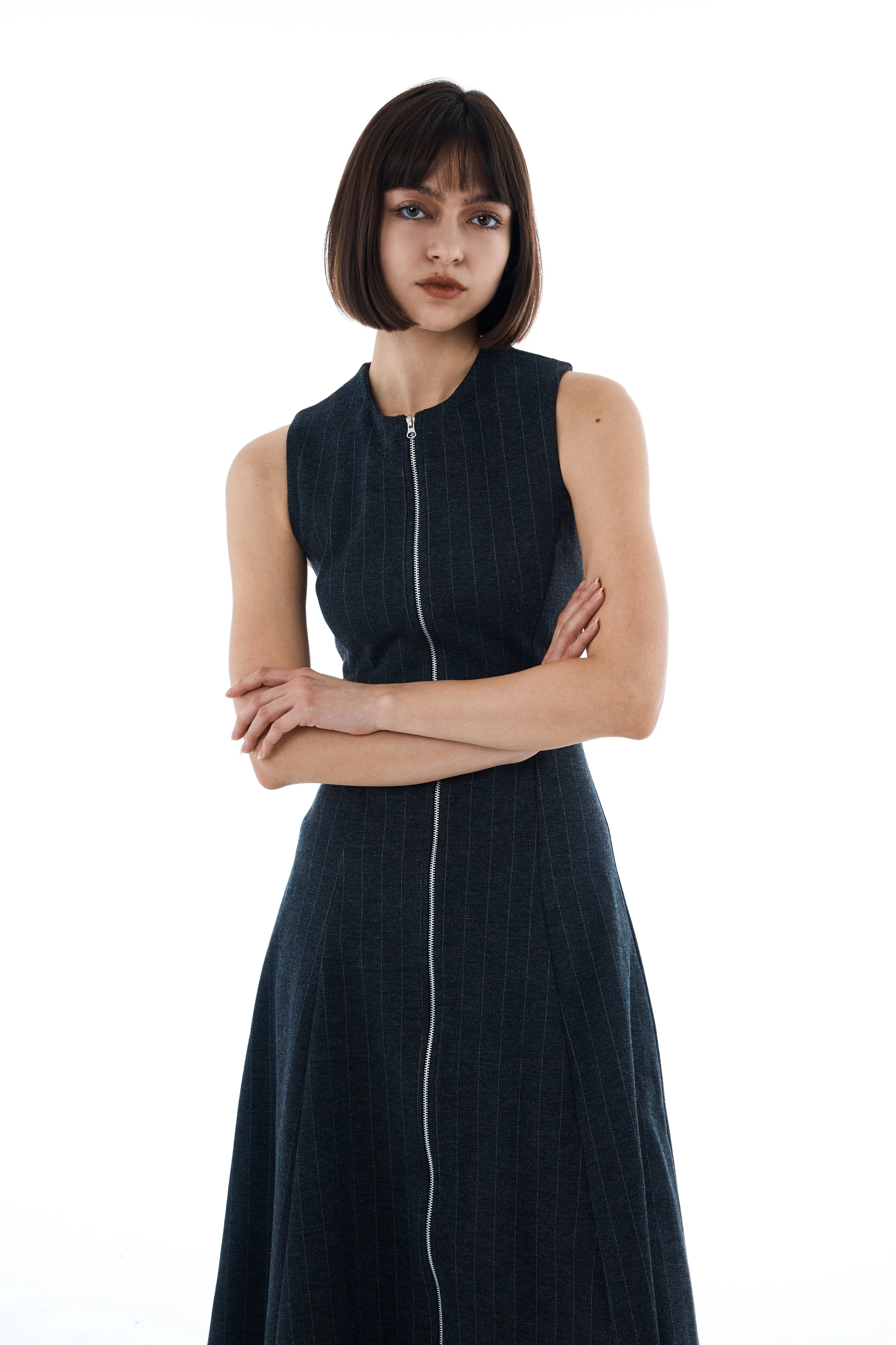 A young woman with shoulder-length dark brown hair and bangs, wearing a sleeveless, dark gray, zip-up dress with vertical pinstripes, standing with arms crossed against a plain white background.