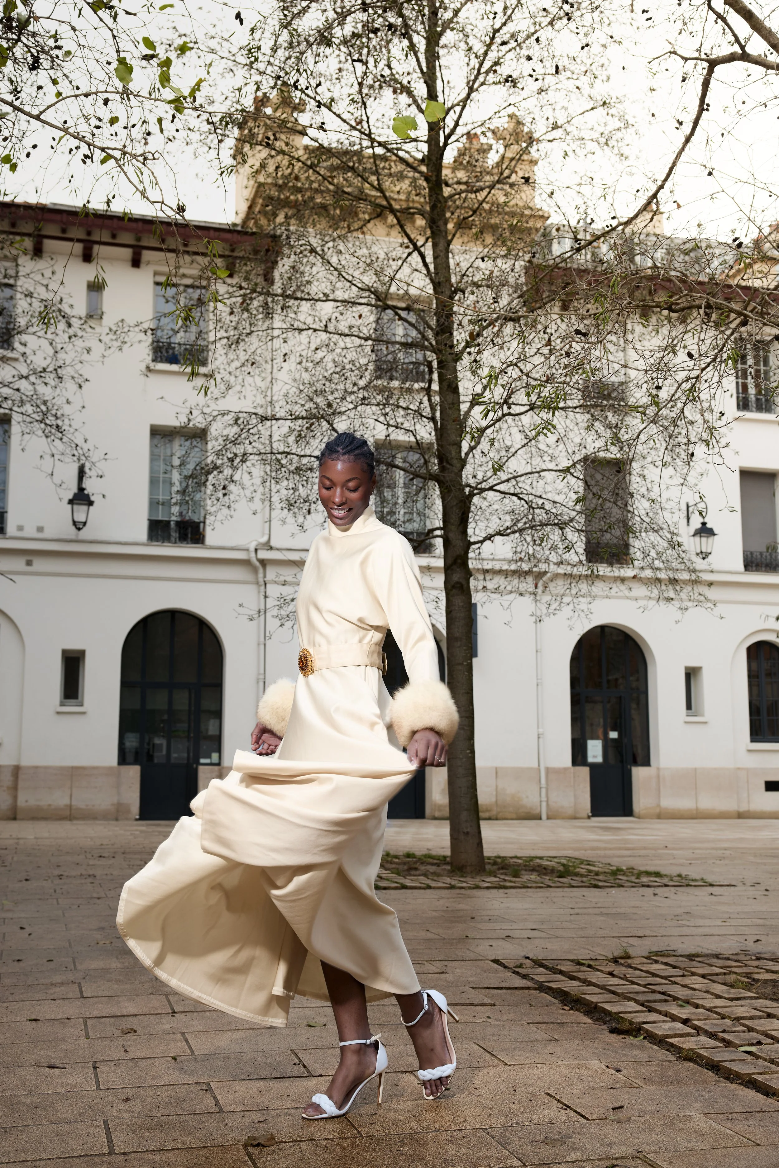 A woman in a cream-colored dress with fur cuffs and high heels twirls on a sidewalk, with a tree and white building in the background.