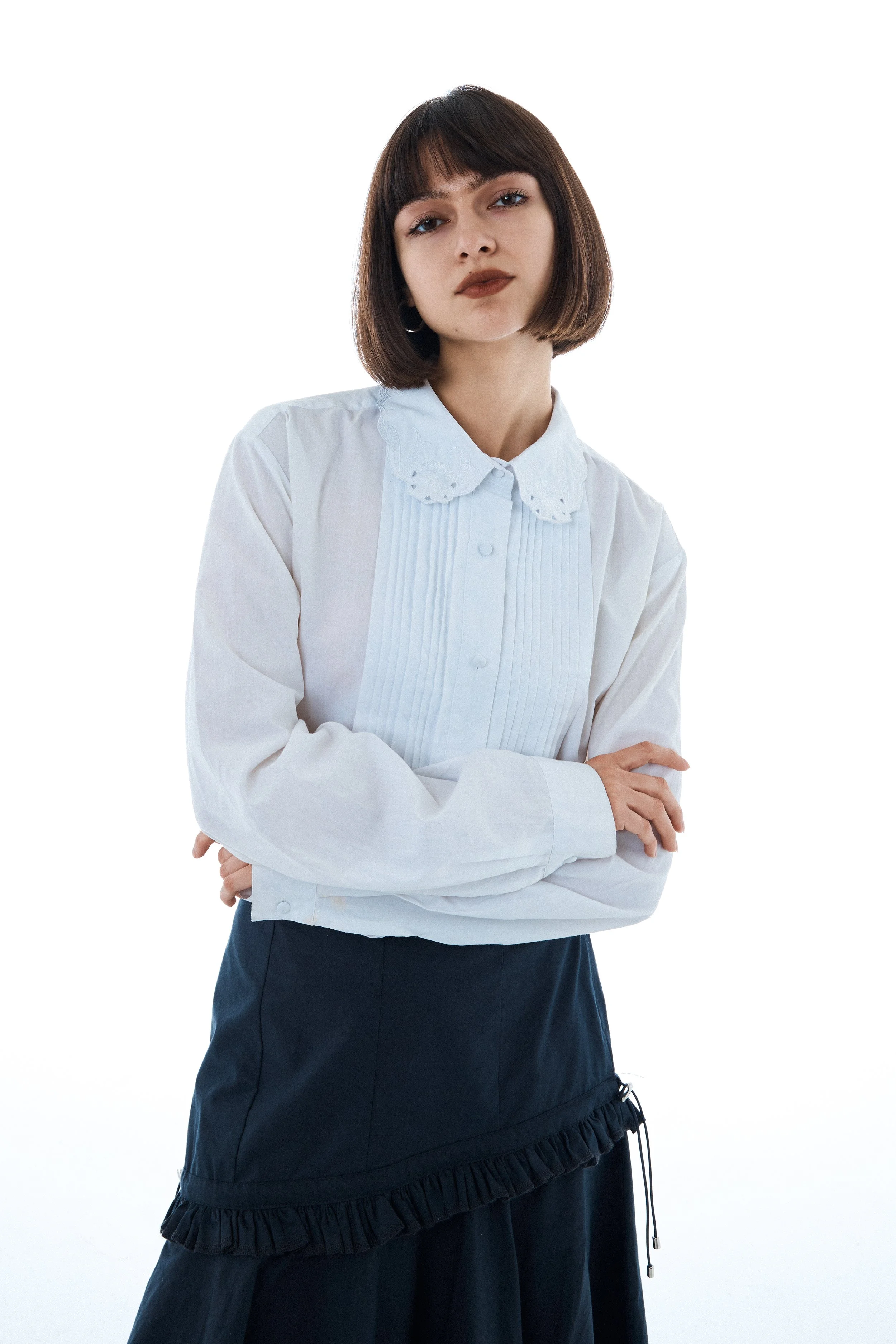 Young woman with dark brown hair and bangs, wearing a white blouse with a Peter Pan collar and pleated front, and a black skirt with ruffle trim, standing with arms crossed against a plain white background.