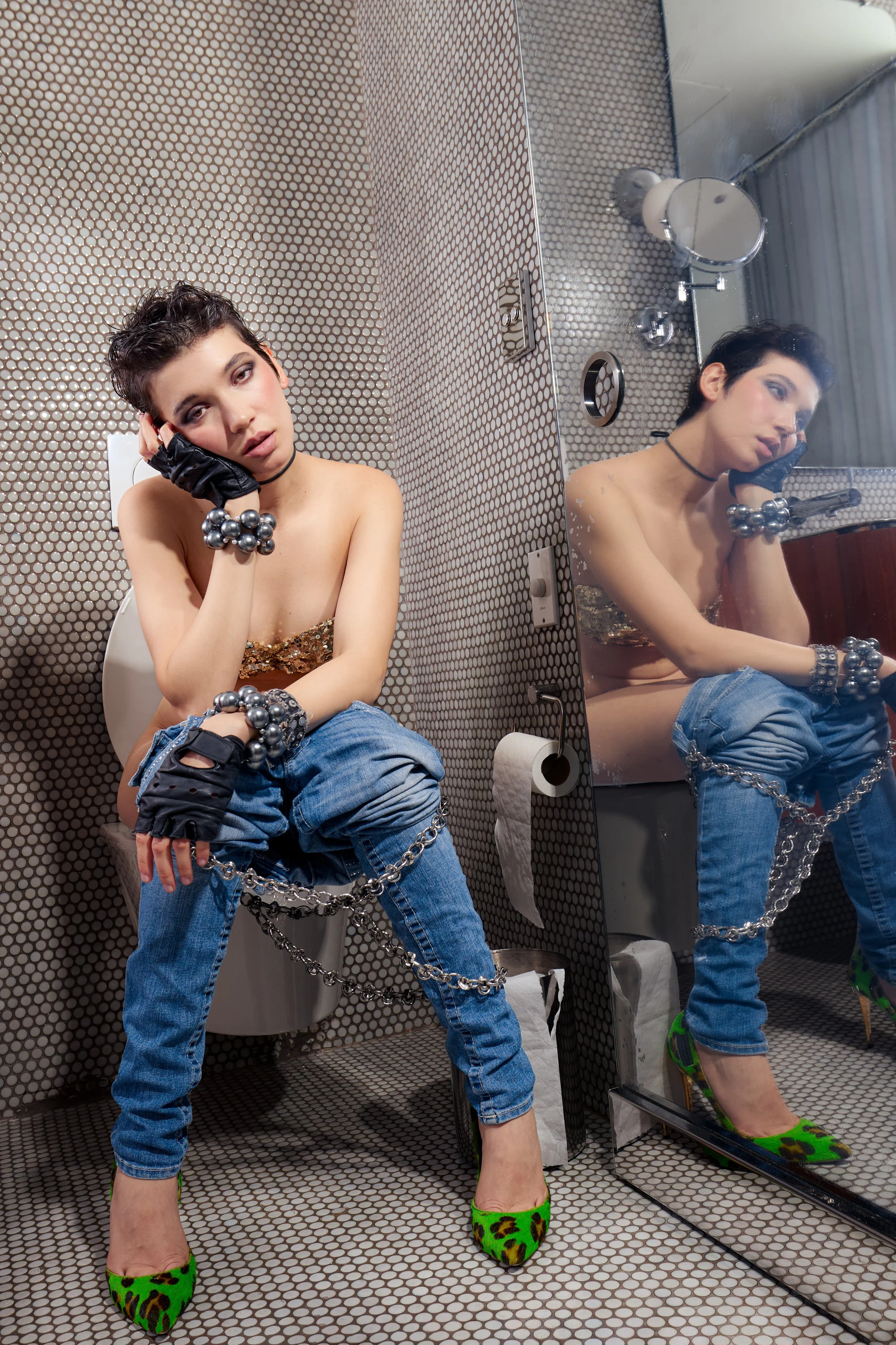 A woman with short dark hair and makeup, sitting on a toilet in a bathroom with small white tiles, wearing a gold top, blue jeans chained at the waist, green leopard print high heels, without pants, black gloves, and chunky silver jewelry, posing for