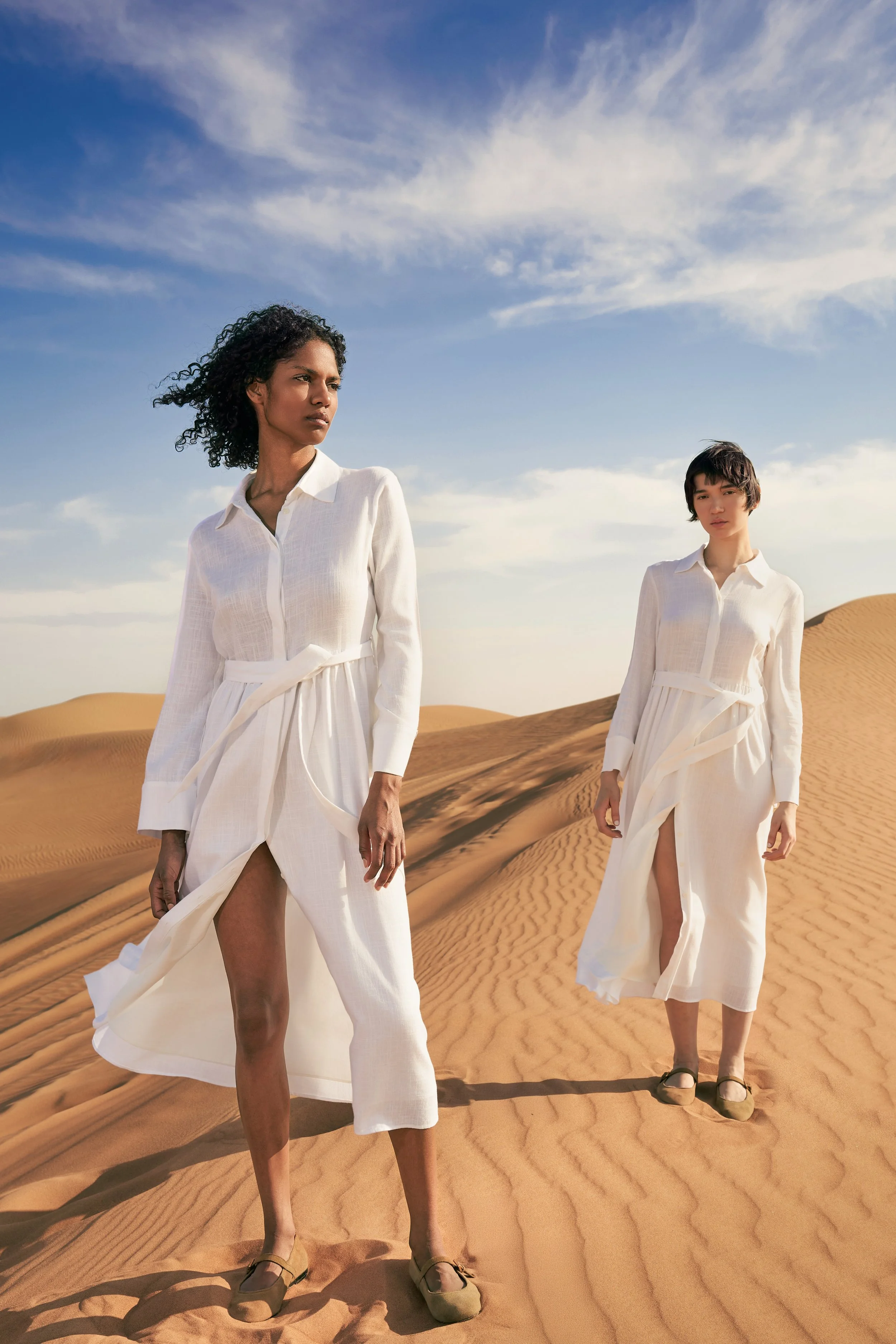 Two women walking on sand dunes in a desert, wearing white long-sleeve dresses with thigh-high slits and beige shoes, under a partly cloudy sky.