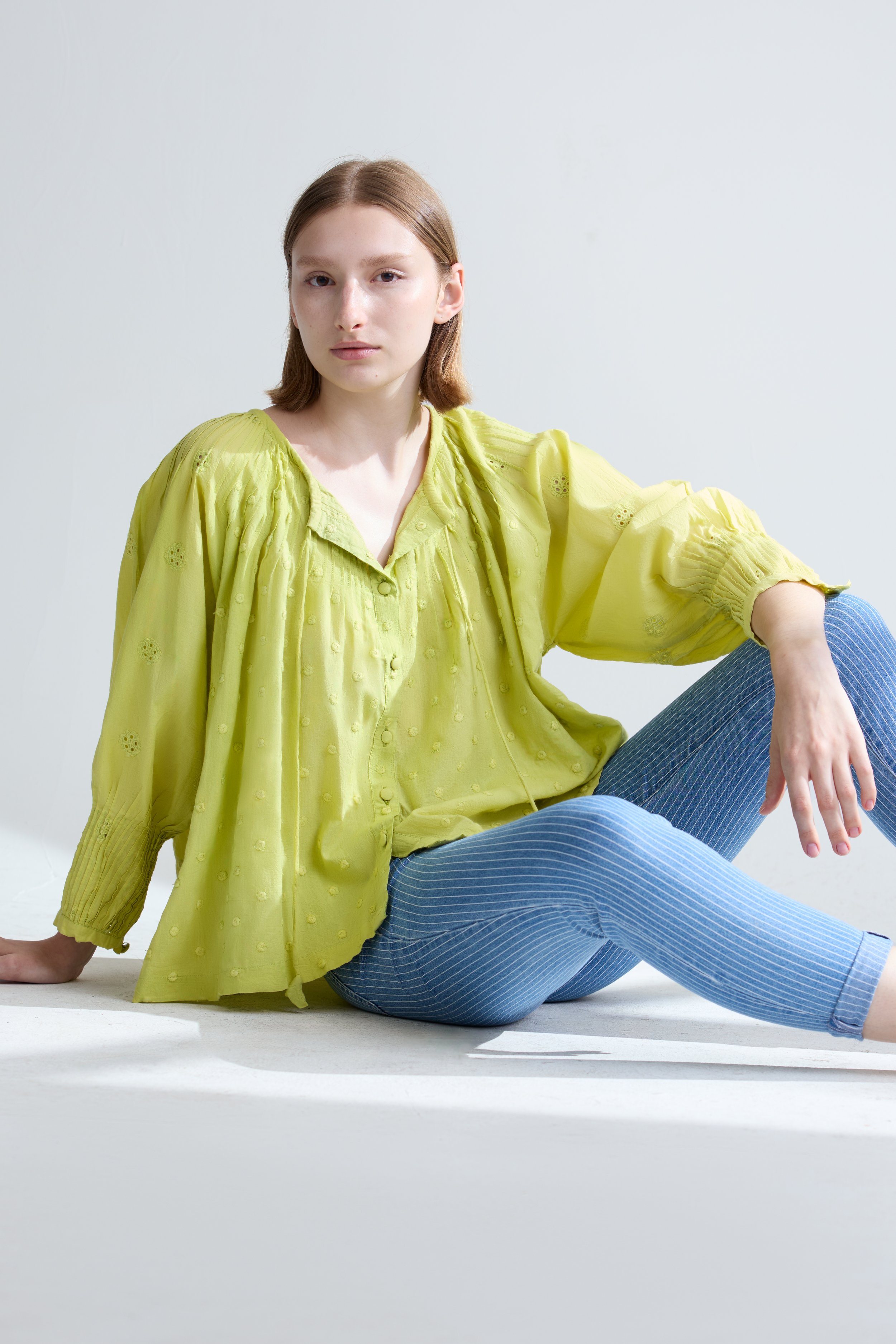 A young woman with shoulder-length hair sits on the floor against a plain white background. She wears a yellow, loose-fitting blouse and blue pinstriped pants, looking directly at the camera with a neutral expression.