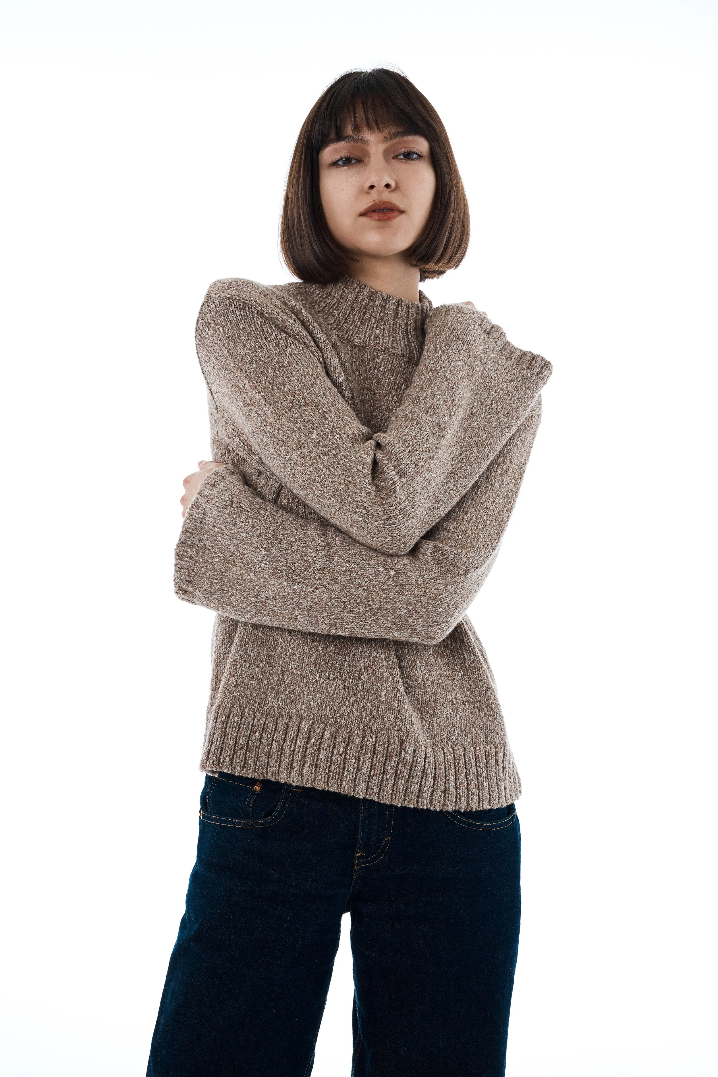 Young woman with short brown hair wearing a beige sweater and dark jeans, standing with arms crossed and looking at the camera.