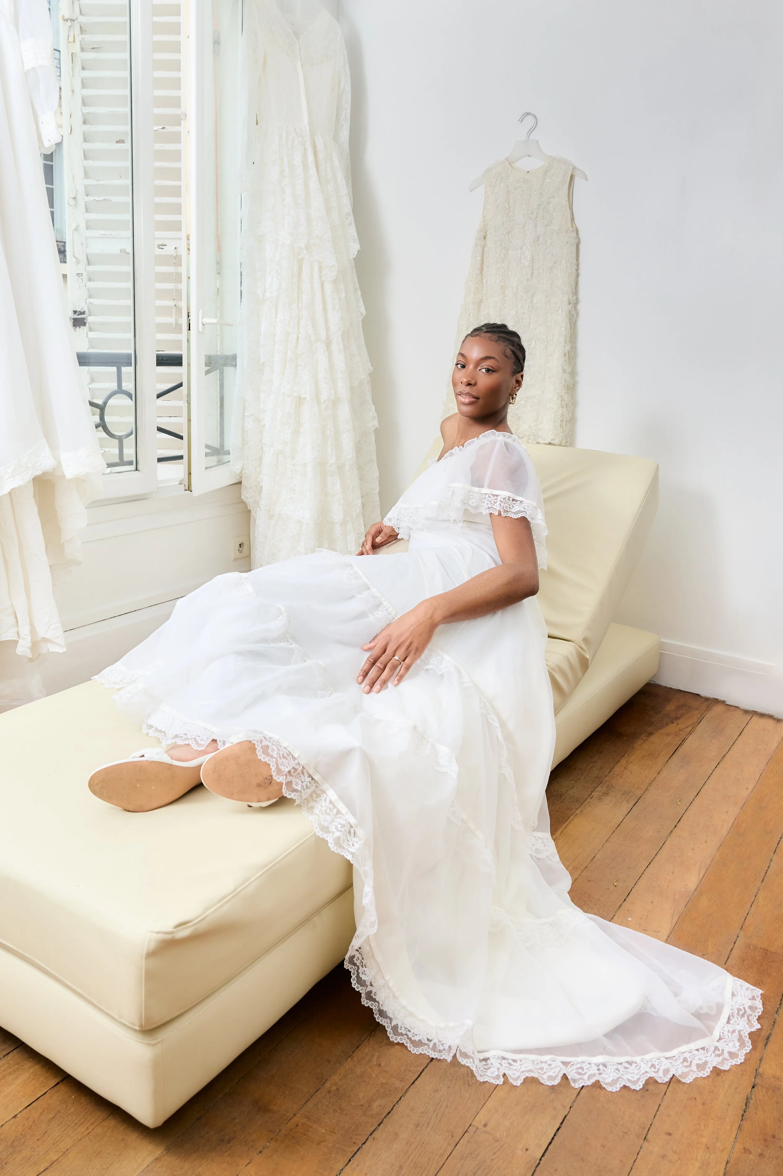 A woman in a white vintage-style dress sitting on a beige chaise lounge, with two other dresses hanging behind her near a window with white shutters.