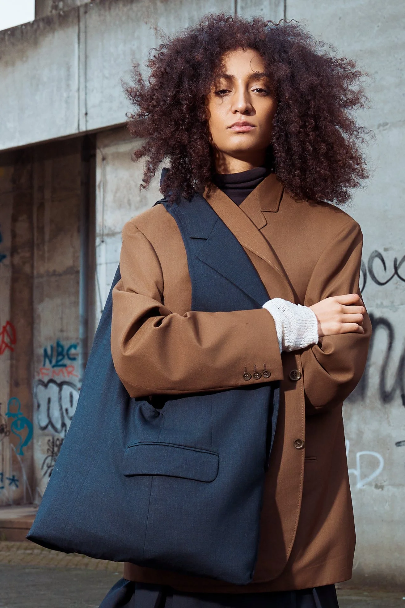 A woman with curly hair wearing a brown blazer, a black turtleneck, a white glove on her left hand, and carrying a large blue shoulder bag stands against a graffiti-covered concrete wall.