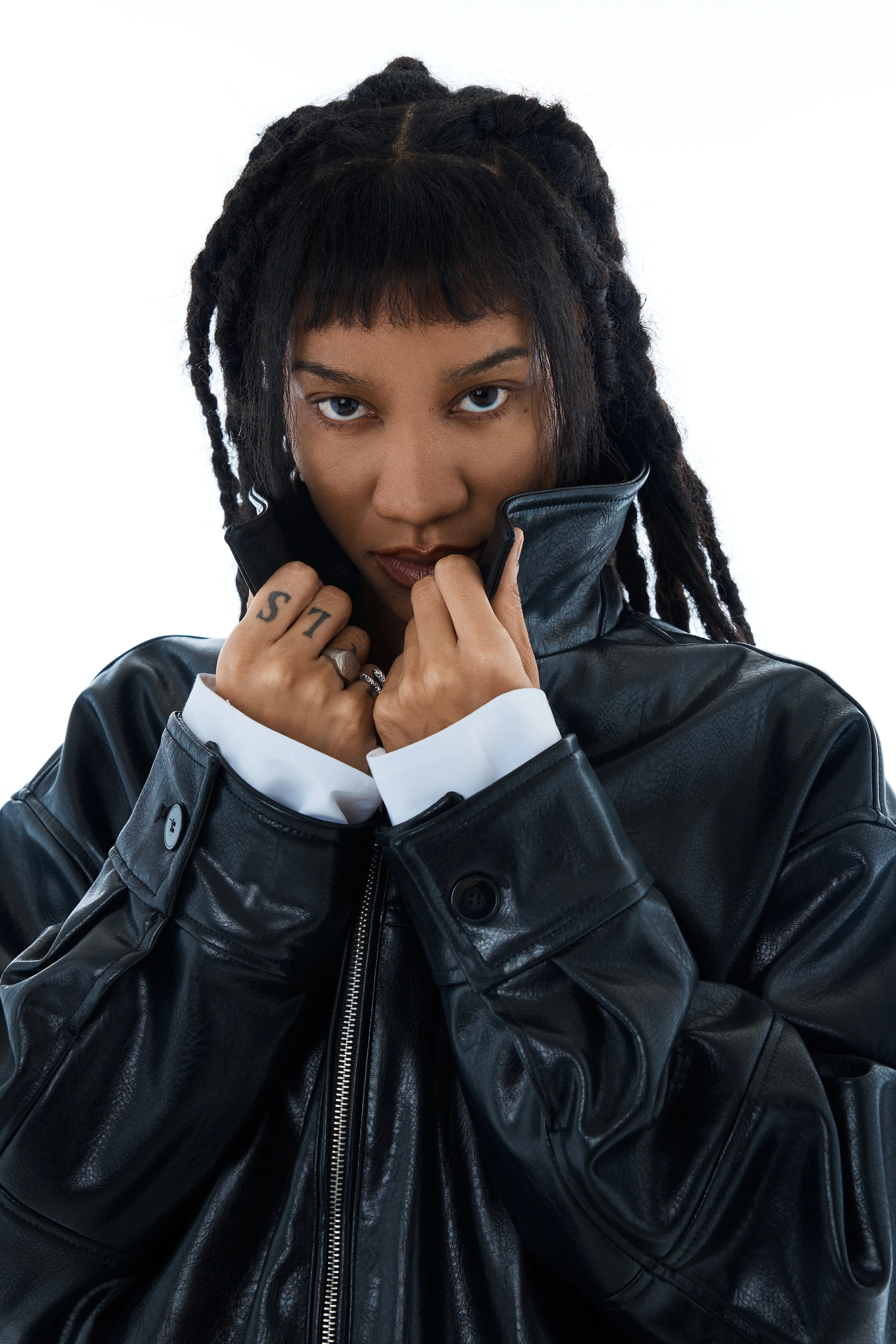 A woman with dreadlocks wearing a black leather jacket with a white shirt underneath, looking directly at the camera and holding the collar of her jacket up.