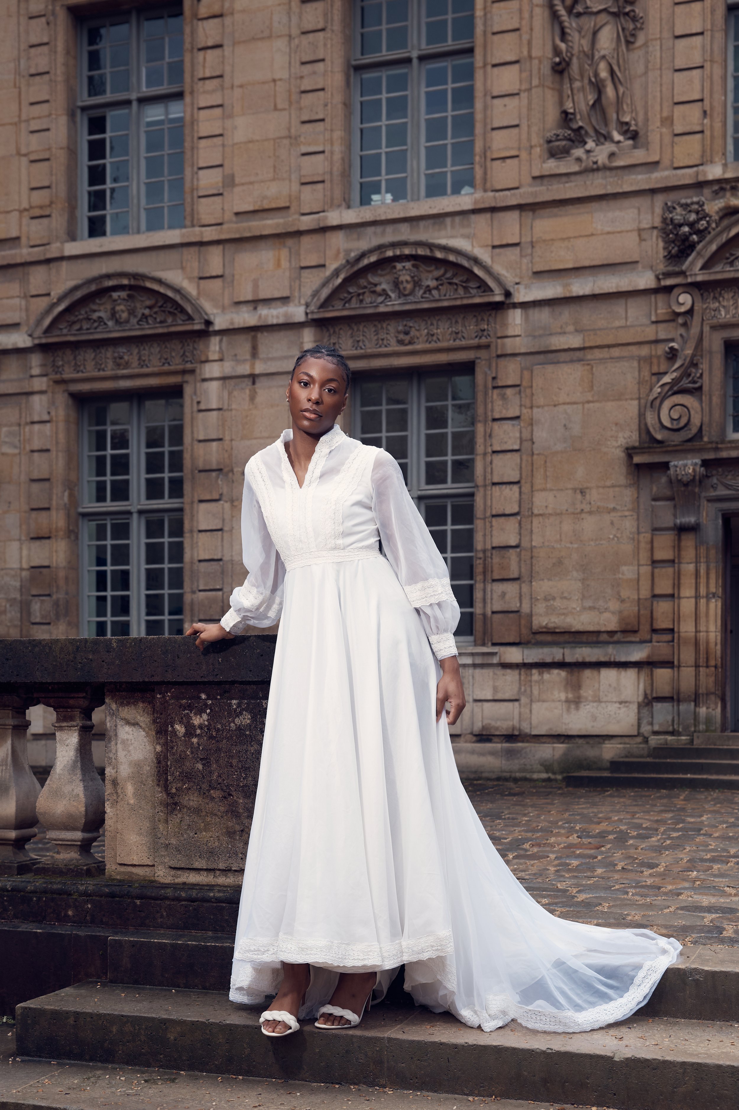 A woman in a white vintage-style wedding dress standing outside in front of a historic stone building.