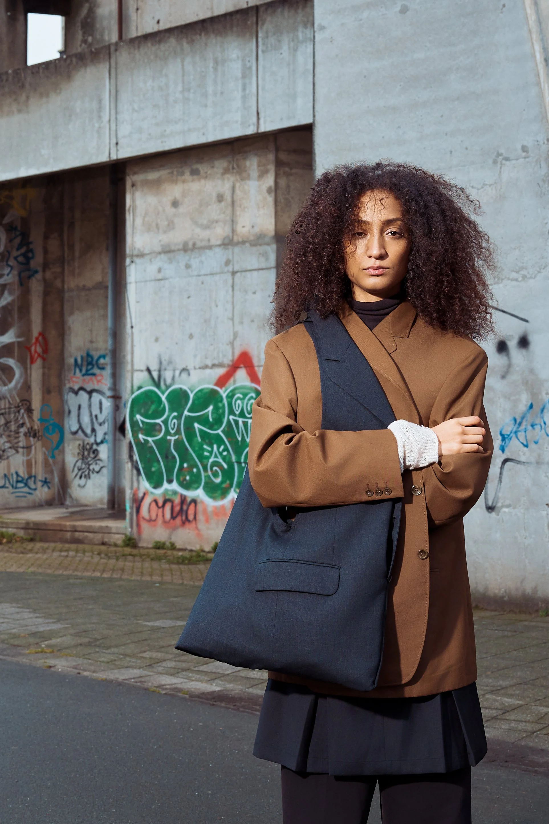 A woman with curly hair standing in front of a graffiti-covered concrete wall, wearing a brown blazer, black pants, and carrying a large black shoulder bag.