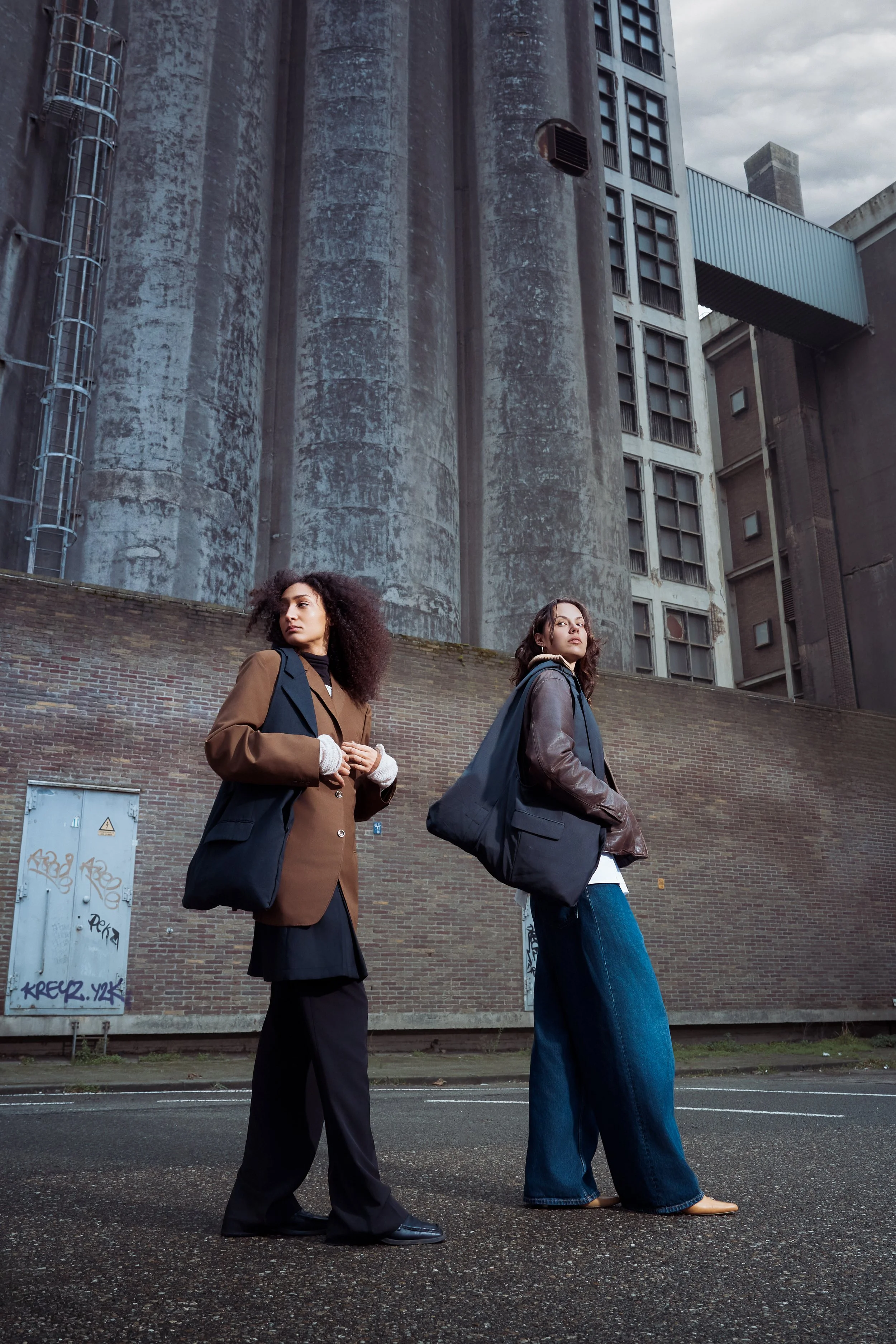Two women standing on a street in front of an industrial building with large silos and graffiti. One woman has curly hair and is wearing a brown and black blazer with wide black pants, and the other has straight hair and is dressed in a brown leather