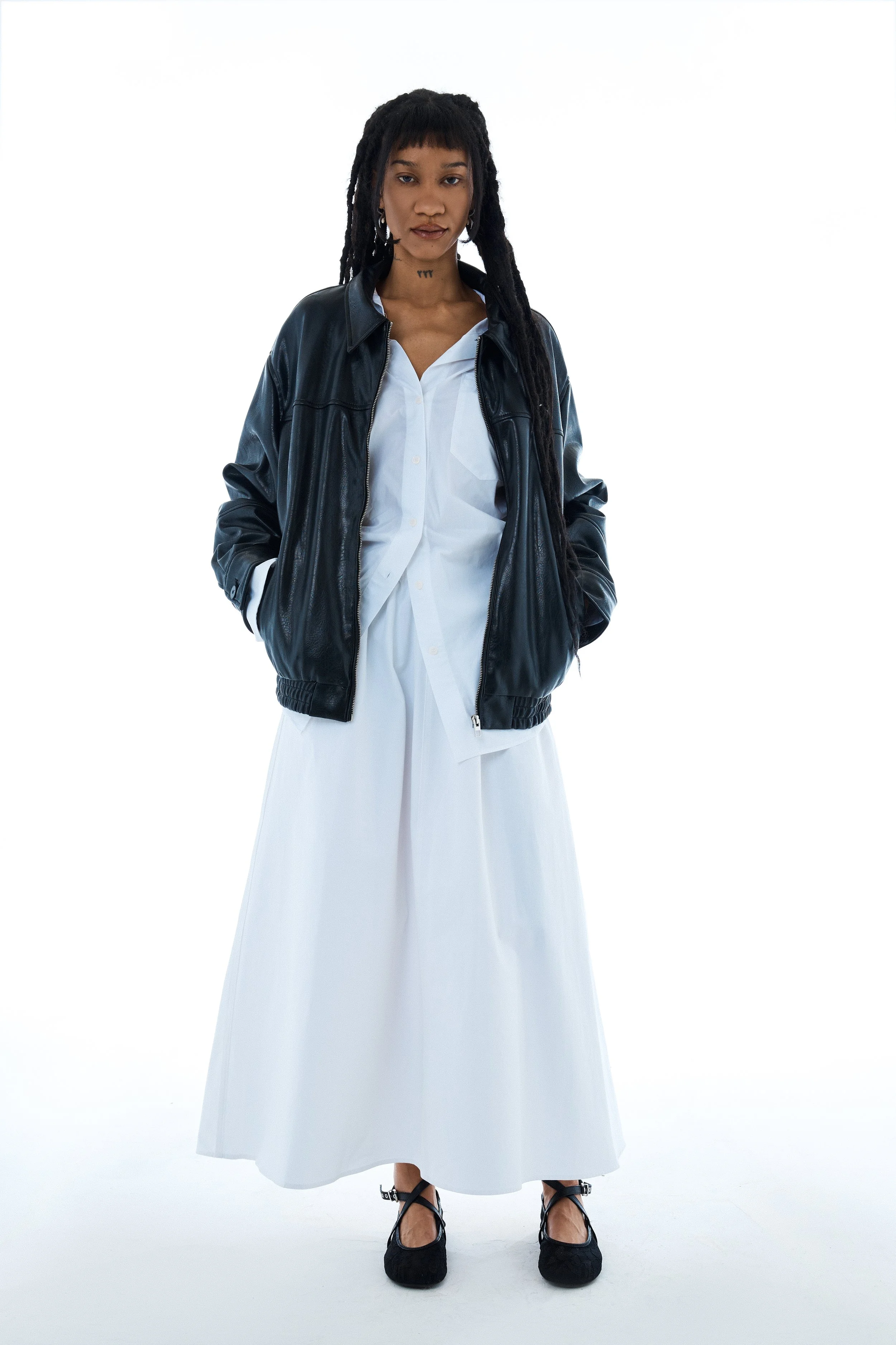 A woman with long dreadlocks wearing a black leather jacket over a white shirt and long white skirt, black shoes, standing against a white background.