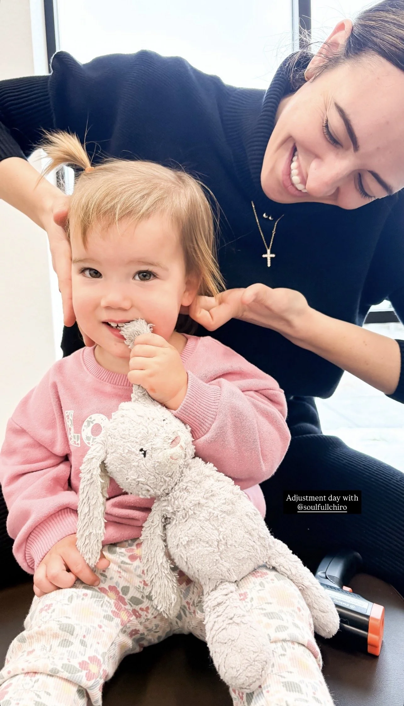 A man and a young girl are on a black chiropractic table. The man is supporting the girl as she lies face down on the table, holding a book or paper. A boy is on the man's back, with his arms around his neck, and appears to be playing or pretending. The room has a gray wall decorated with pictures, a mirror, and a cartoon dinosaur painting in the background.