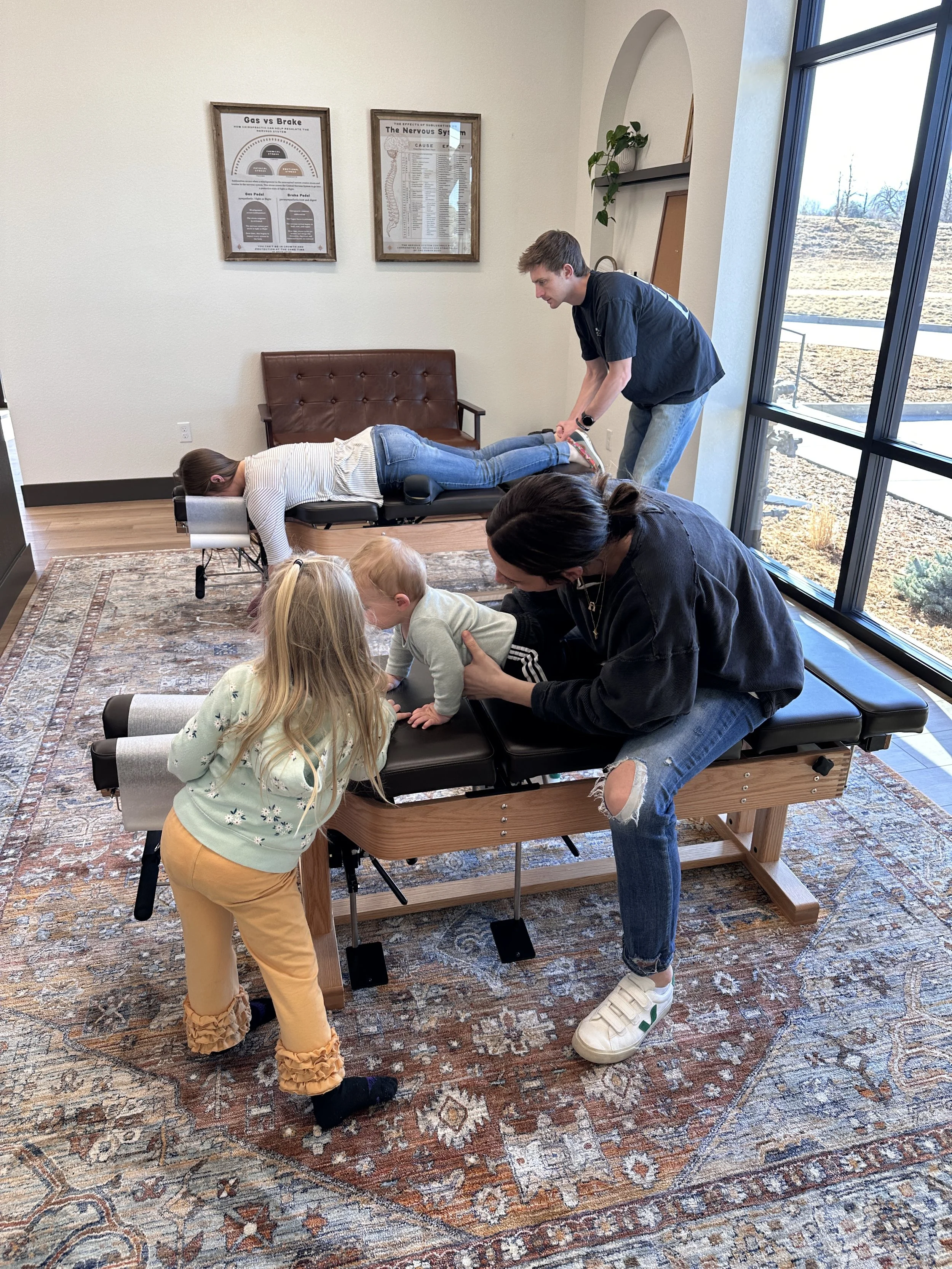 A woman receiving a chiropractic adjustment from a female chiropractor while two young children sit on her lap and one sits on the chiropractic table, in a clinic room with framed posters on the wall.