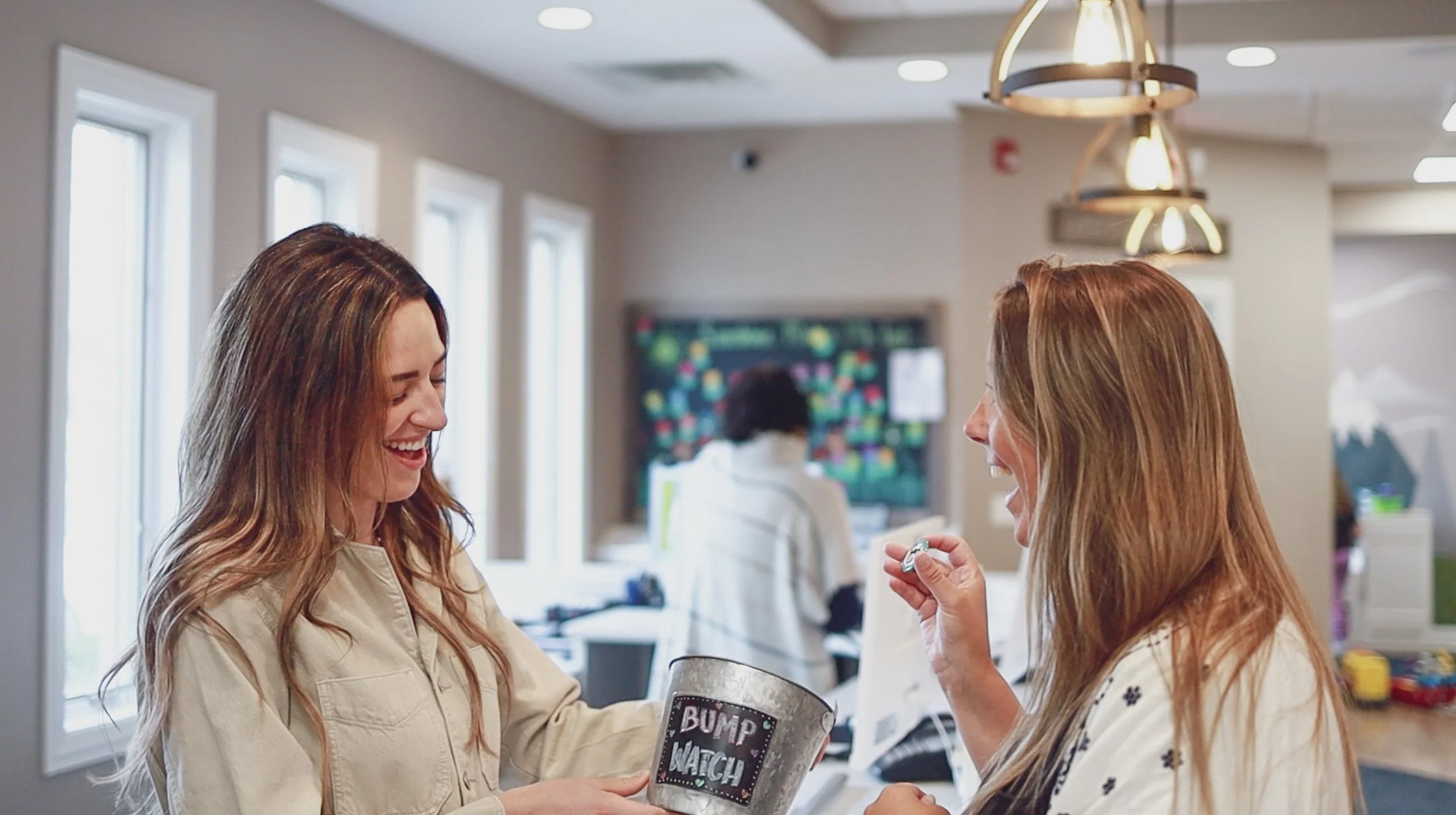 Two women smiling and laughing in a brightly lit indoor setting, one holding a container labeled 'Bump Watch,' engaging in a cheerful conversation about becoming pregnancy under perinatal chiropractic care.