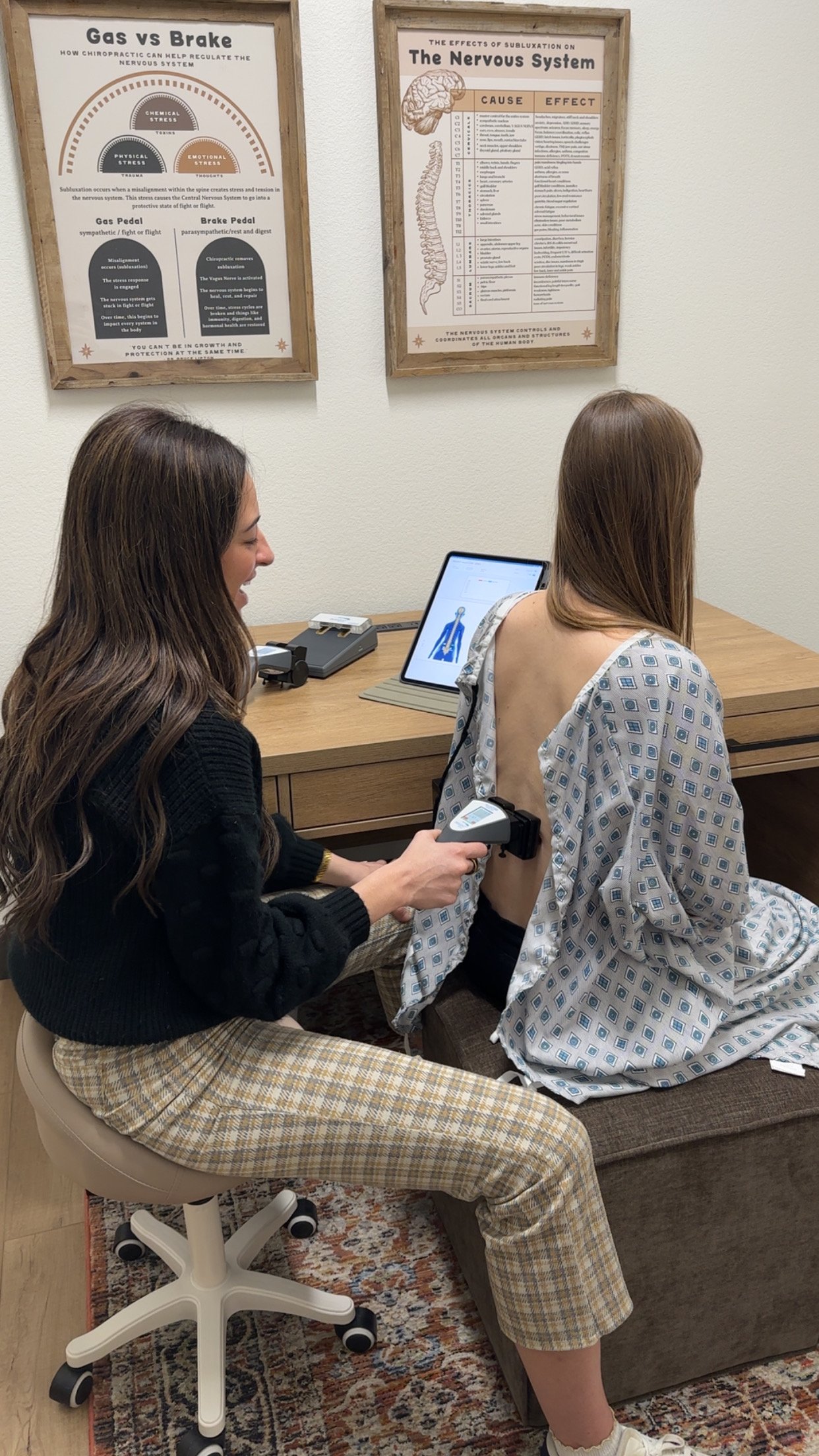 Woman giving a neck massage to a woman lying down in a treatment room with neutral gray walls, wooden floor, framed bulletin board, and decorative wall shelves in the background.