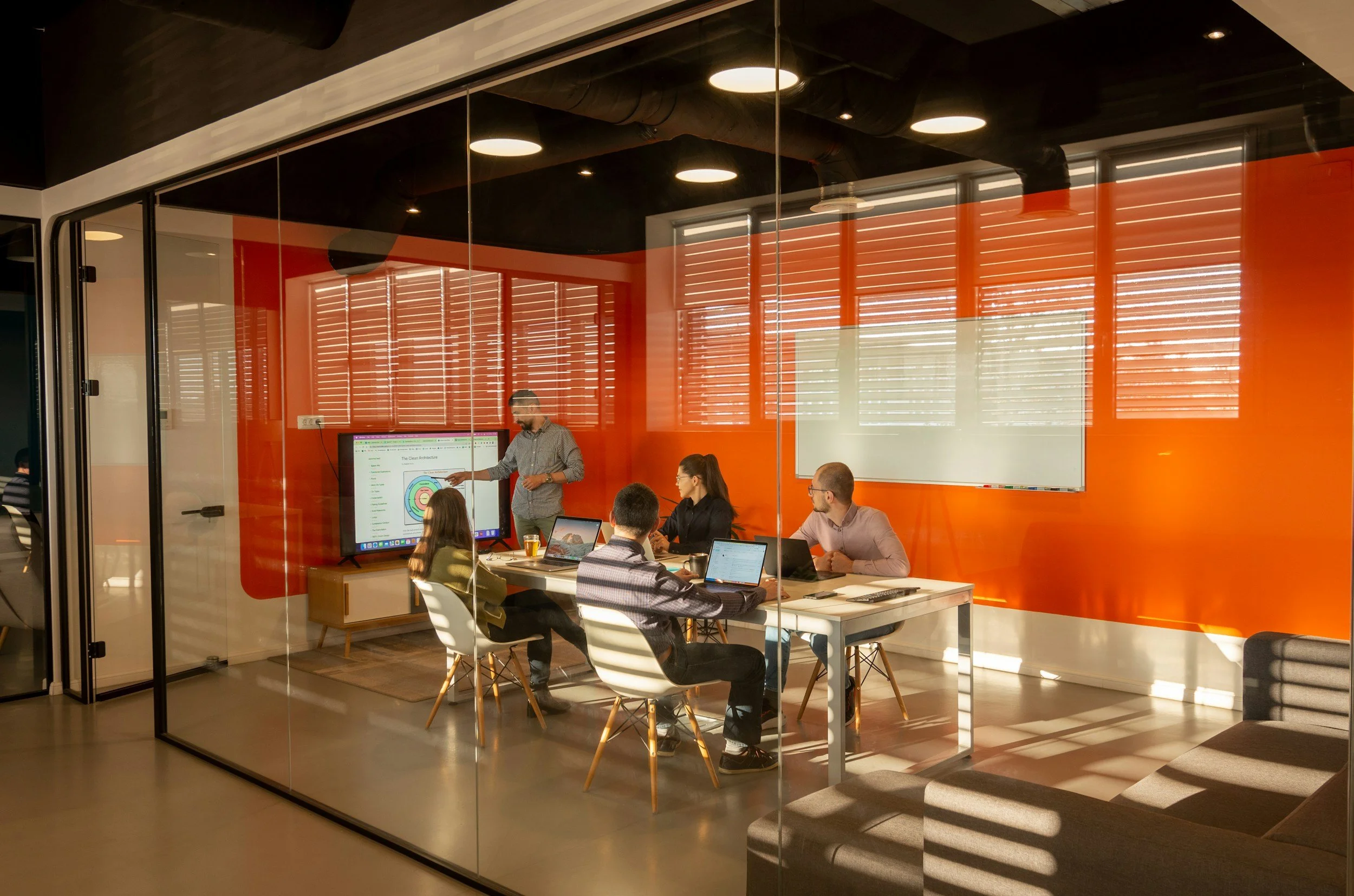 A modern conference room with five people sitting around a white table, facing a man who is standing and presenting slides on a large screen. The room has large windows with horizontal blinds that let in sunlight, and a whiteboard on the orange wall behind the speaker.