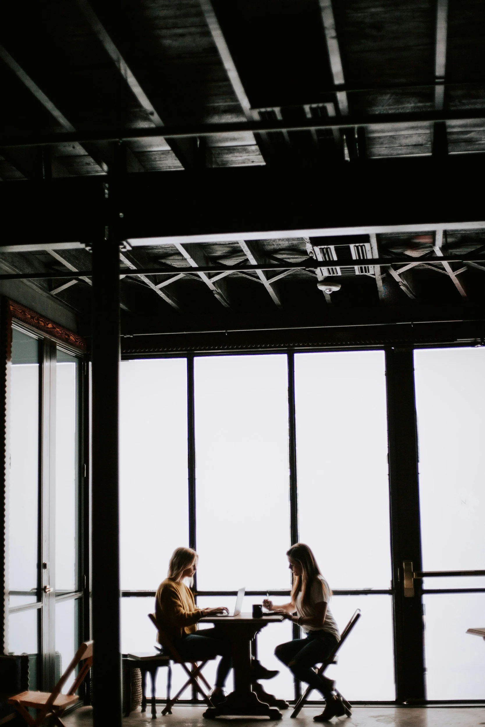 Two women sit at a small round table by large windows in a modern indoor space, working on a laptop and writing, with minimal interior decor and a dark ceiling.