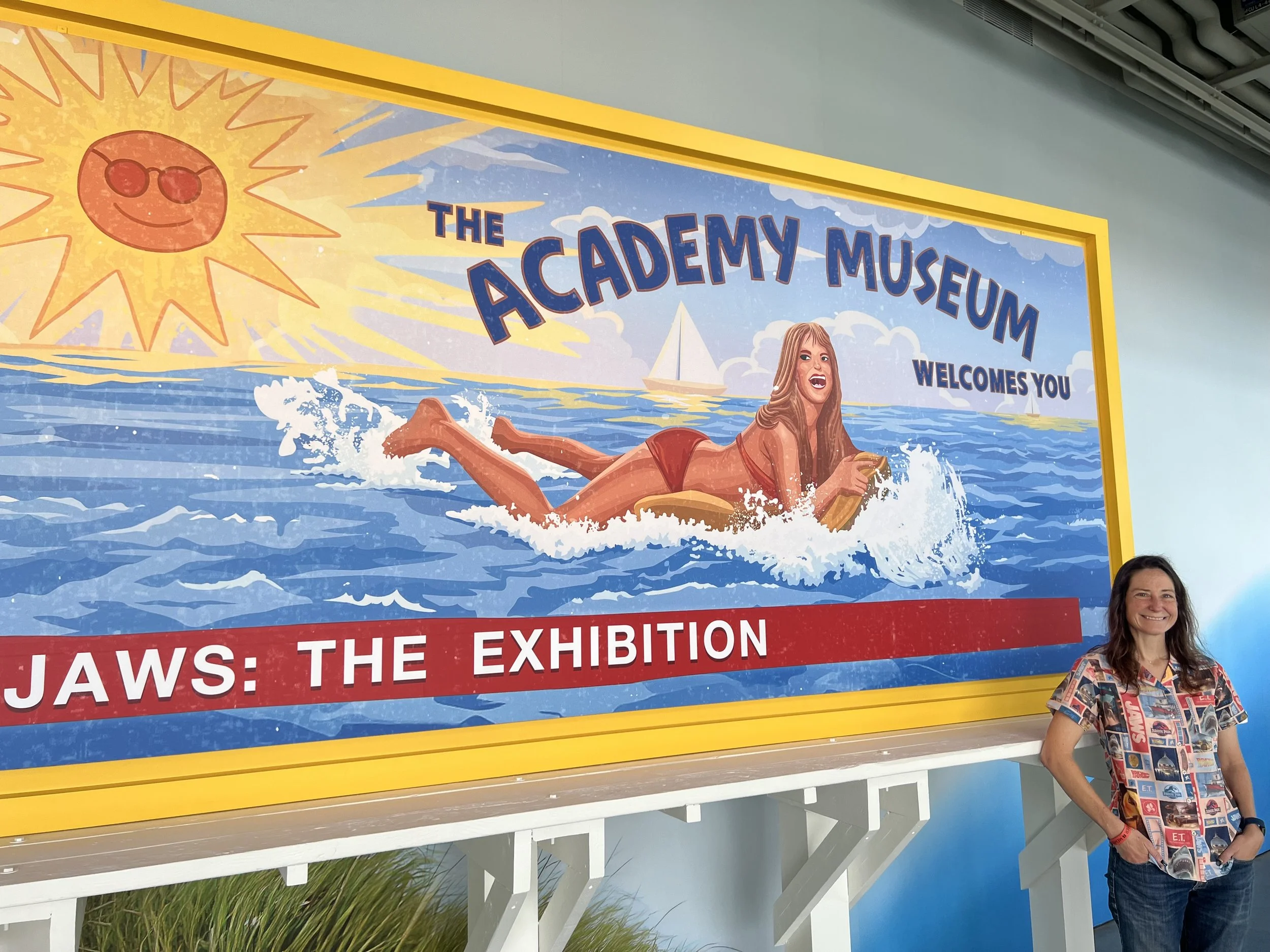 The Amity Island billboard from Jaws, with a happy woman on a floaty in the ocean and a smiling sun, with the text "The Academy Museum Welcomes You!"  This is before the shark attack, obviously.