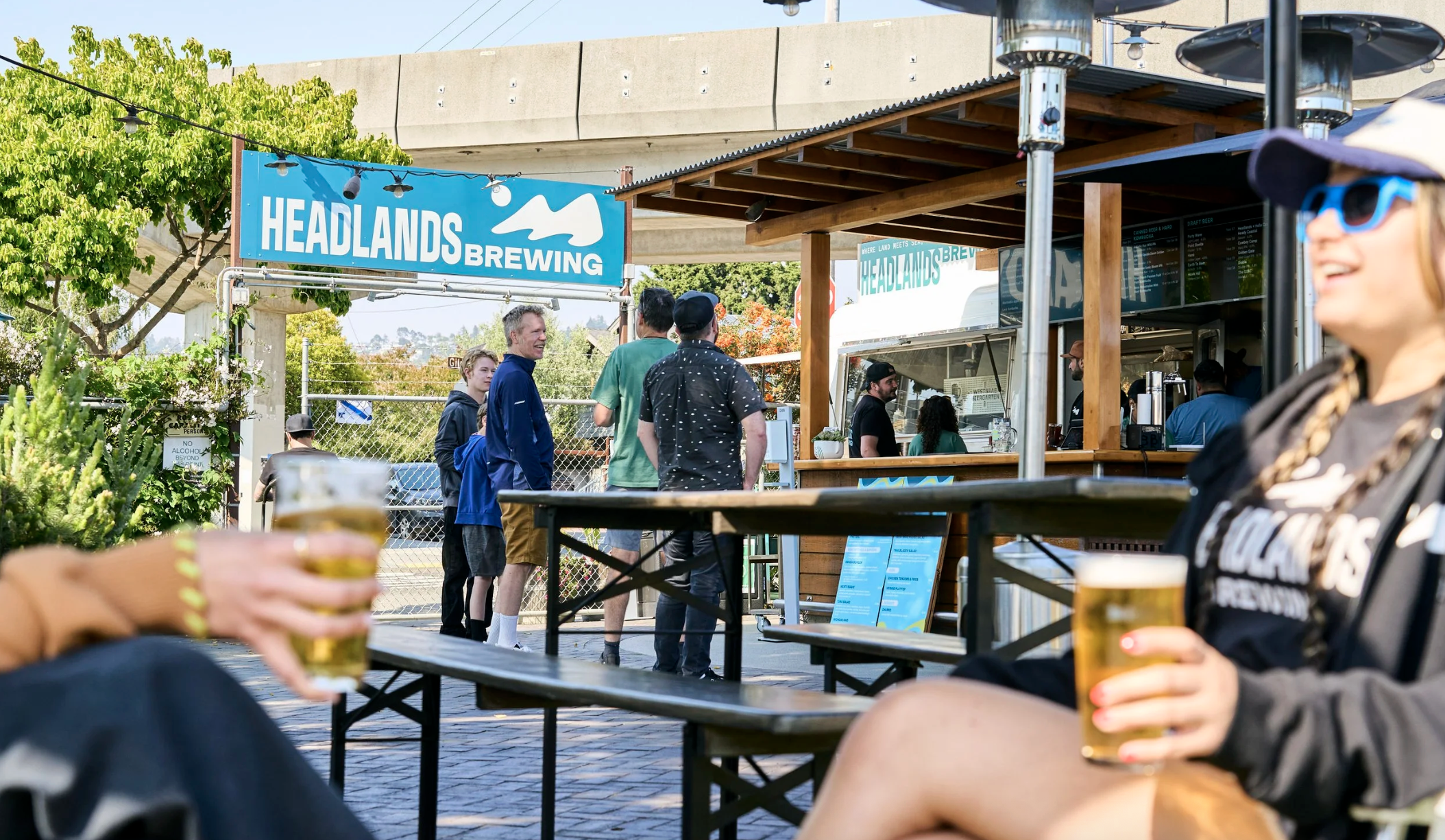 People enjoying outdoor drinks at Headlands Brewing, a brewery with a blue sign, with customers ordering at the counter in the background, and outdoor seating with wooden tables.